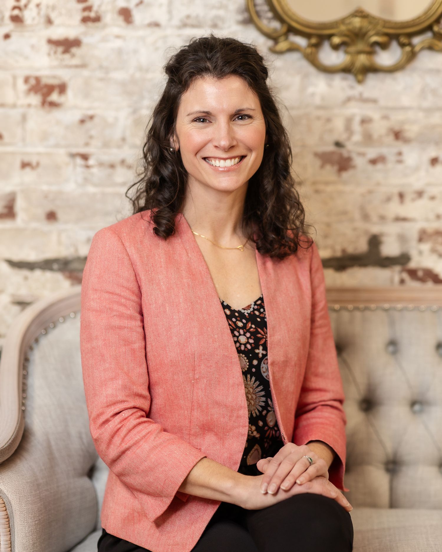 Smiling woman in pink blazer sitting on elegant chair against rustic brick wall backdrop.