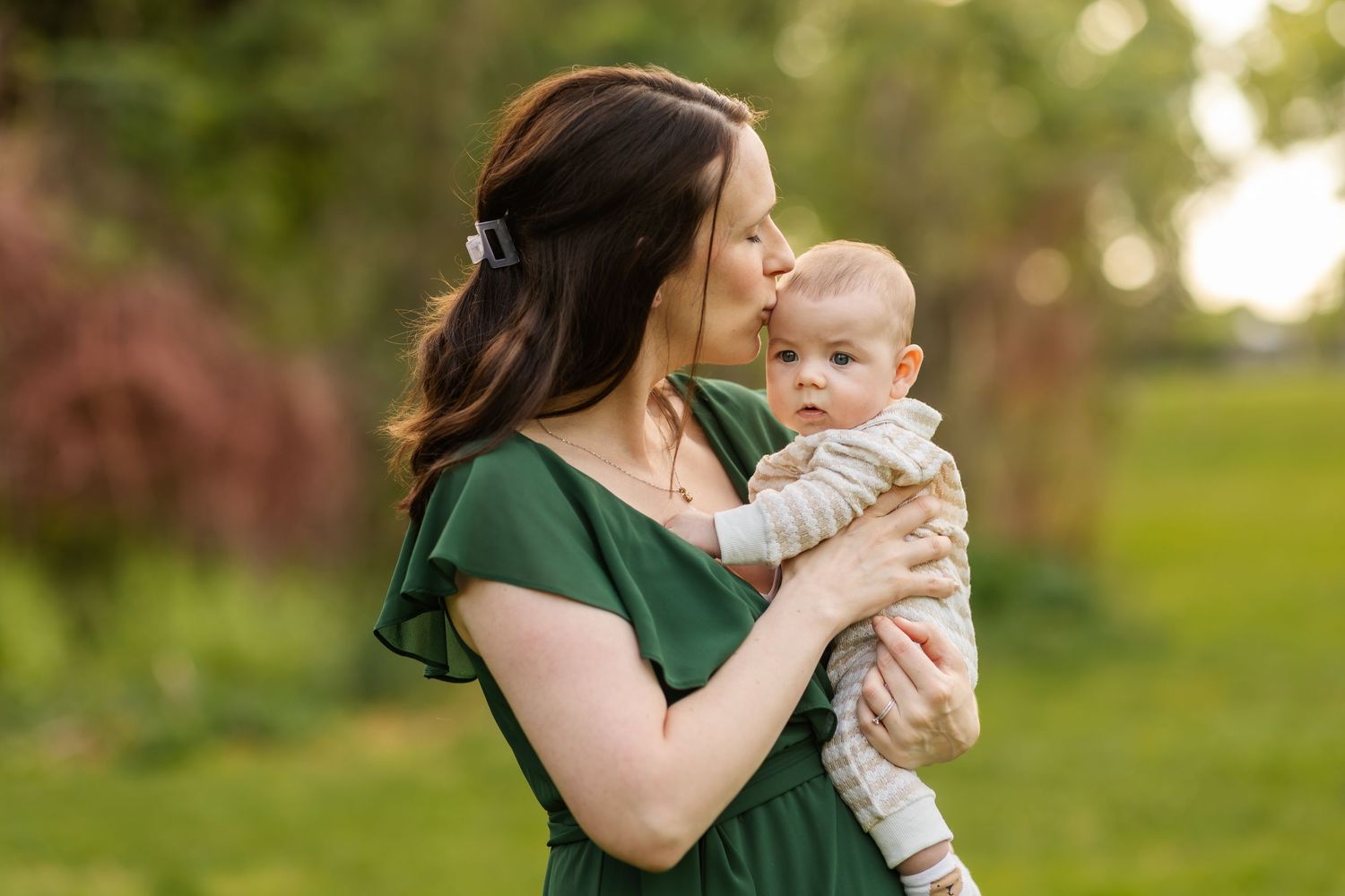 A mother in a green dress tenderly kisses her baby while standing outdoors in a lush green park.