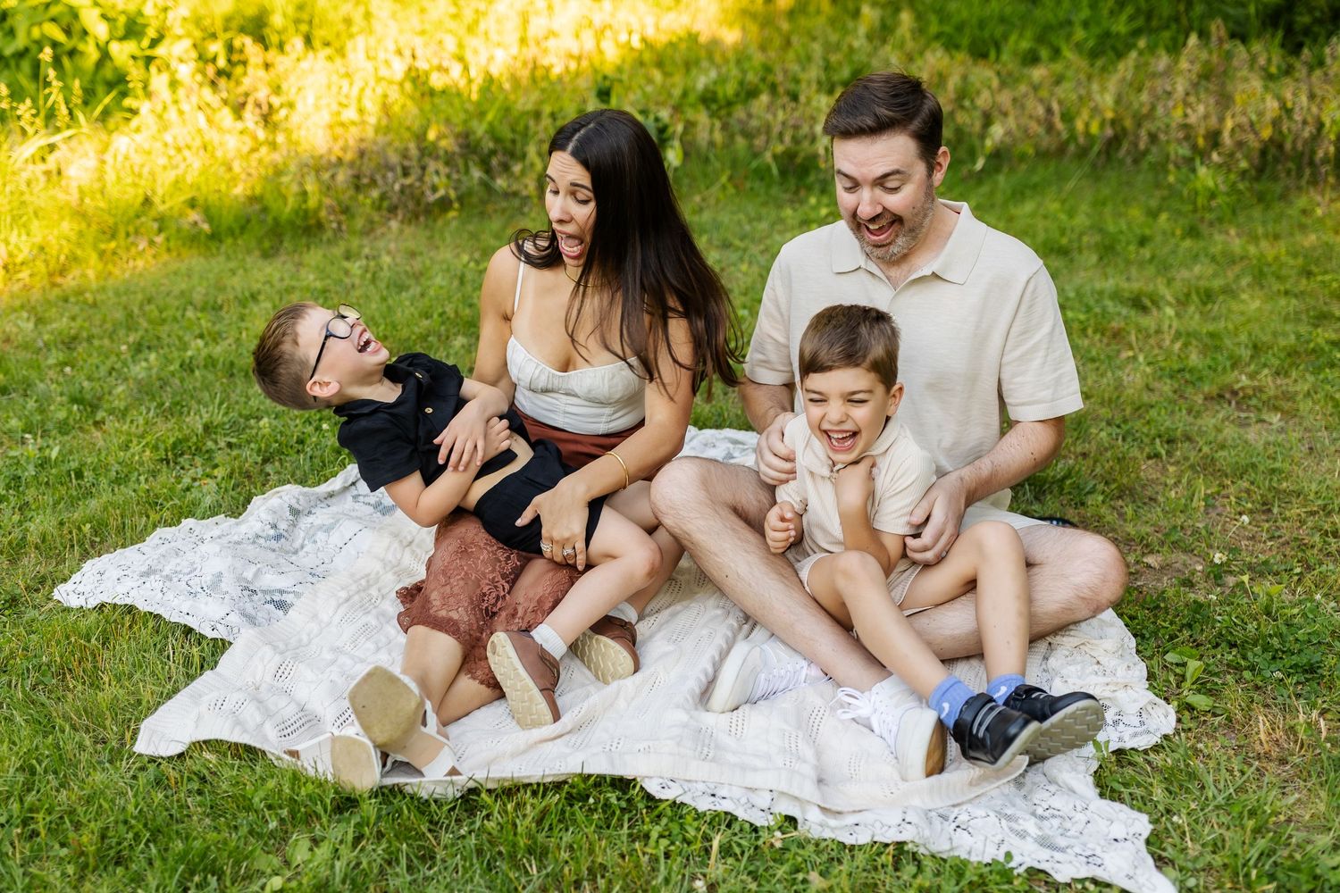 Happy family of four sitting on white blanket in sunny park, parents laughing with two young boys outdoors.
