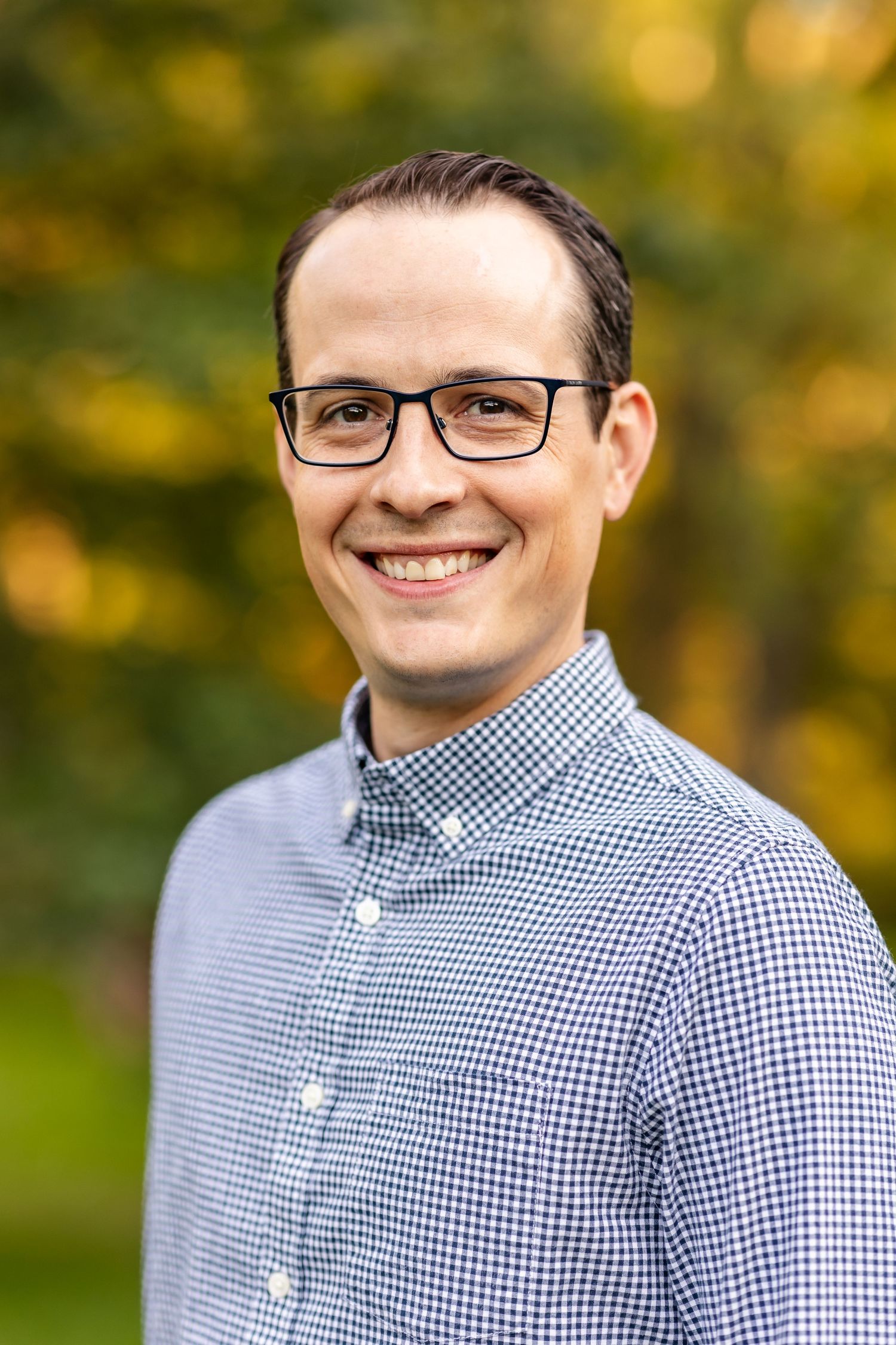 Cheerful professional headshot in blue gingham shirt outdoors with fall foliage.