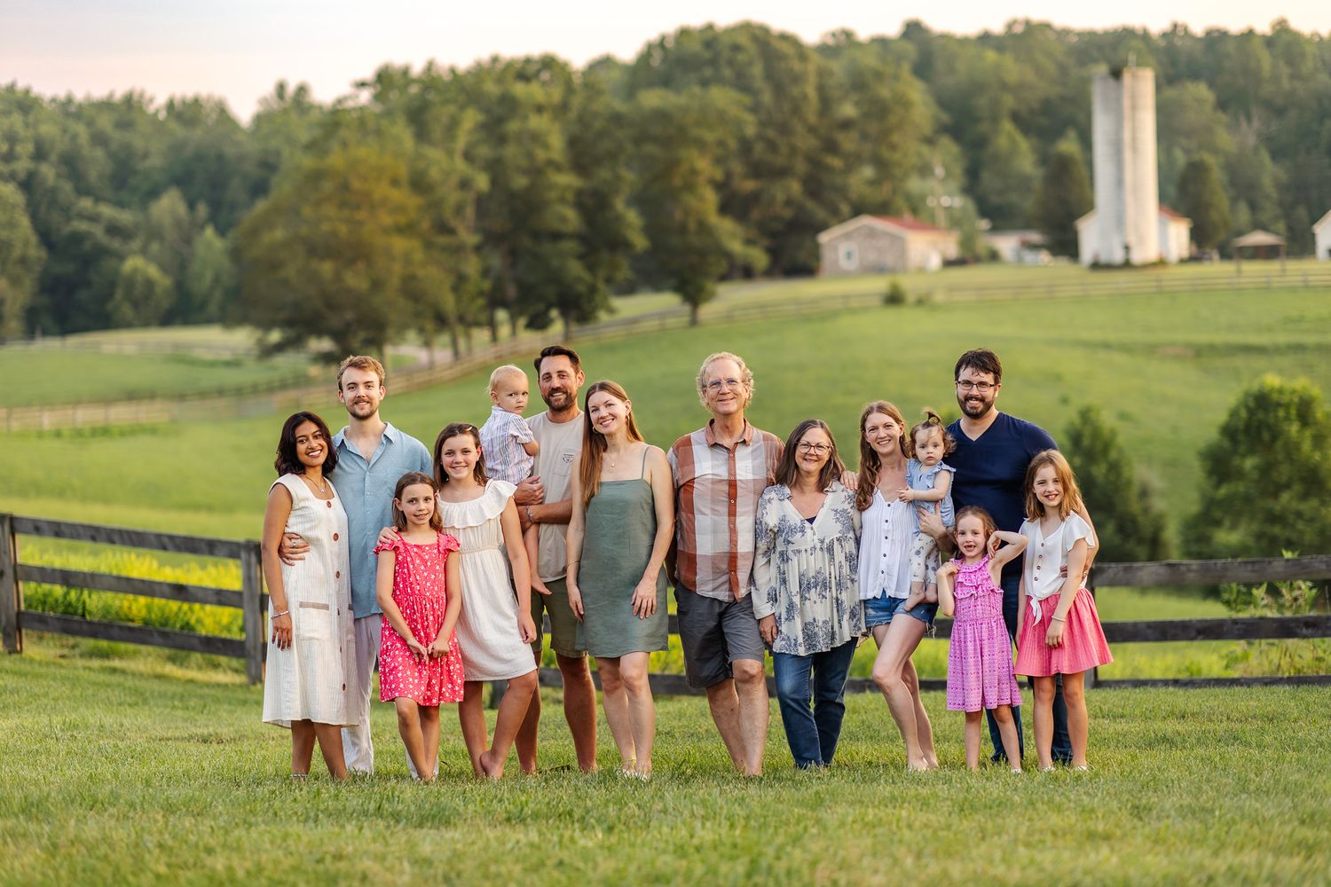 Large multigenerational family smiling together on a lush green farm field at golden hour with a barn in the background.