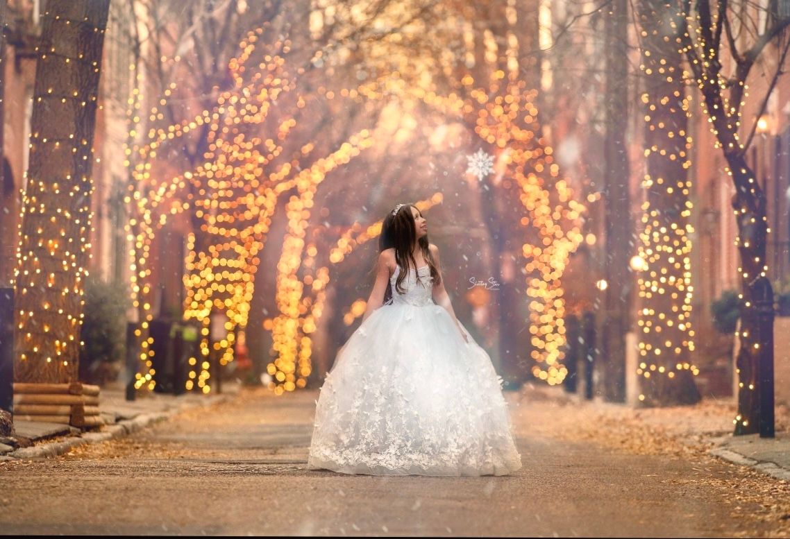 Girl wearing puffy white ballgown stands in the street and gazes at the twinkling lights on the trees around her