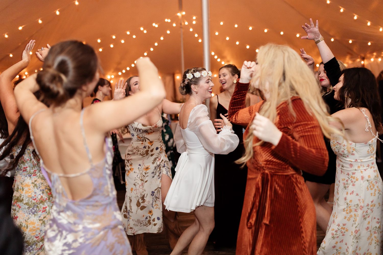 Wedding reception guests dance and celebrate on a dimly lit dance floor decorated with string lights overhead.
