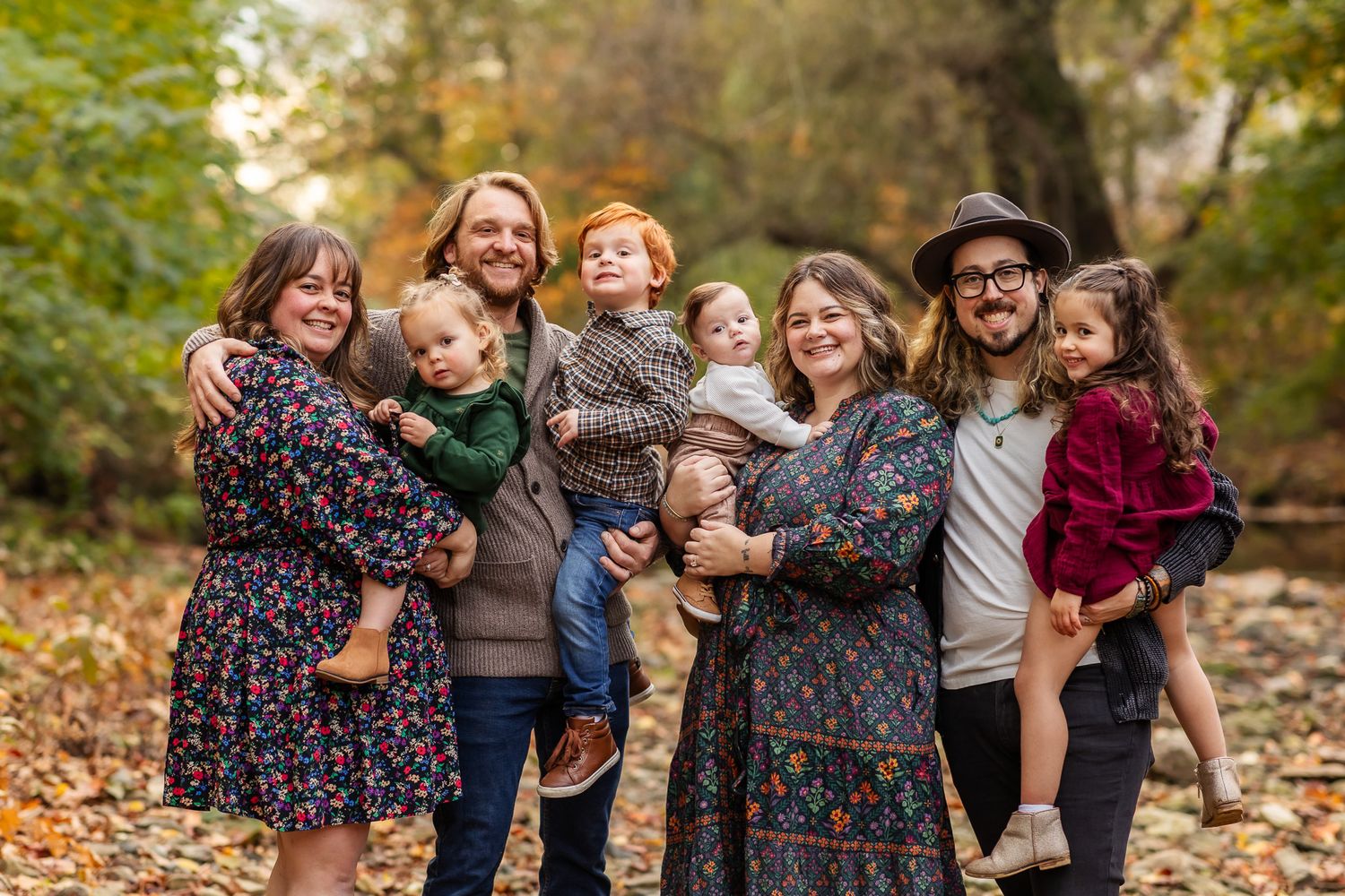 Family of seven smiling outdoors in autumn forest setting with colorful fall foliage in background.
