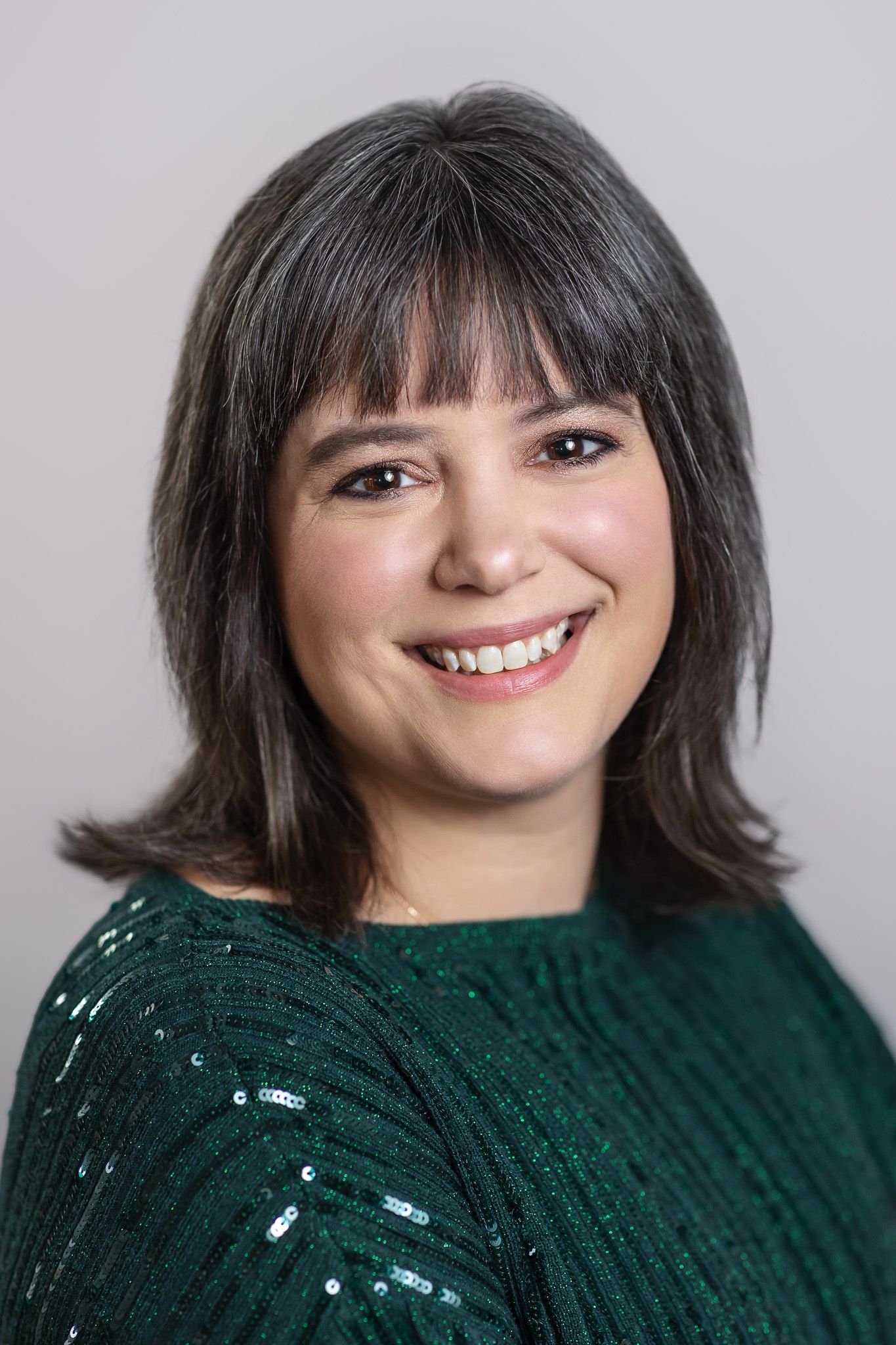 Professional headshot of someone wearing a sparkly green sweater with dark layered hair and bangs against a light background.