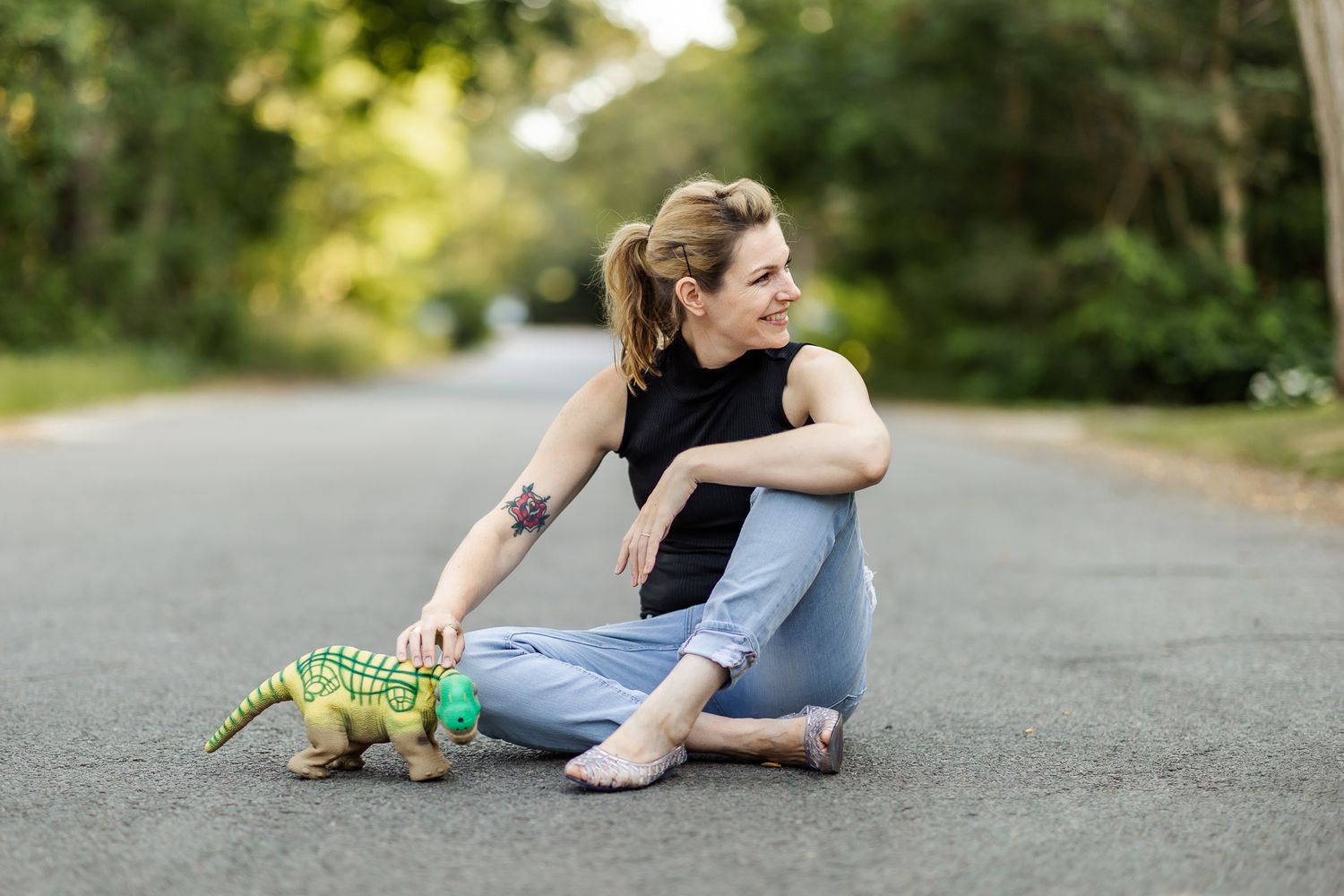 Kate Darling sits in the road petting dinosaur robot and smiling off to the side