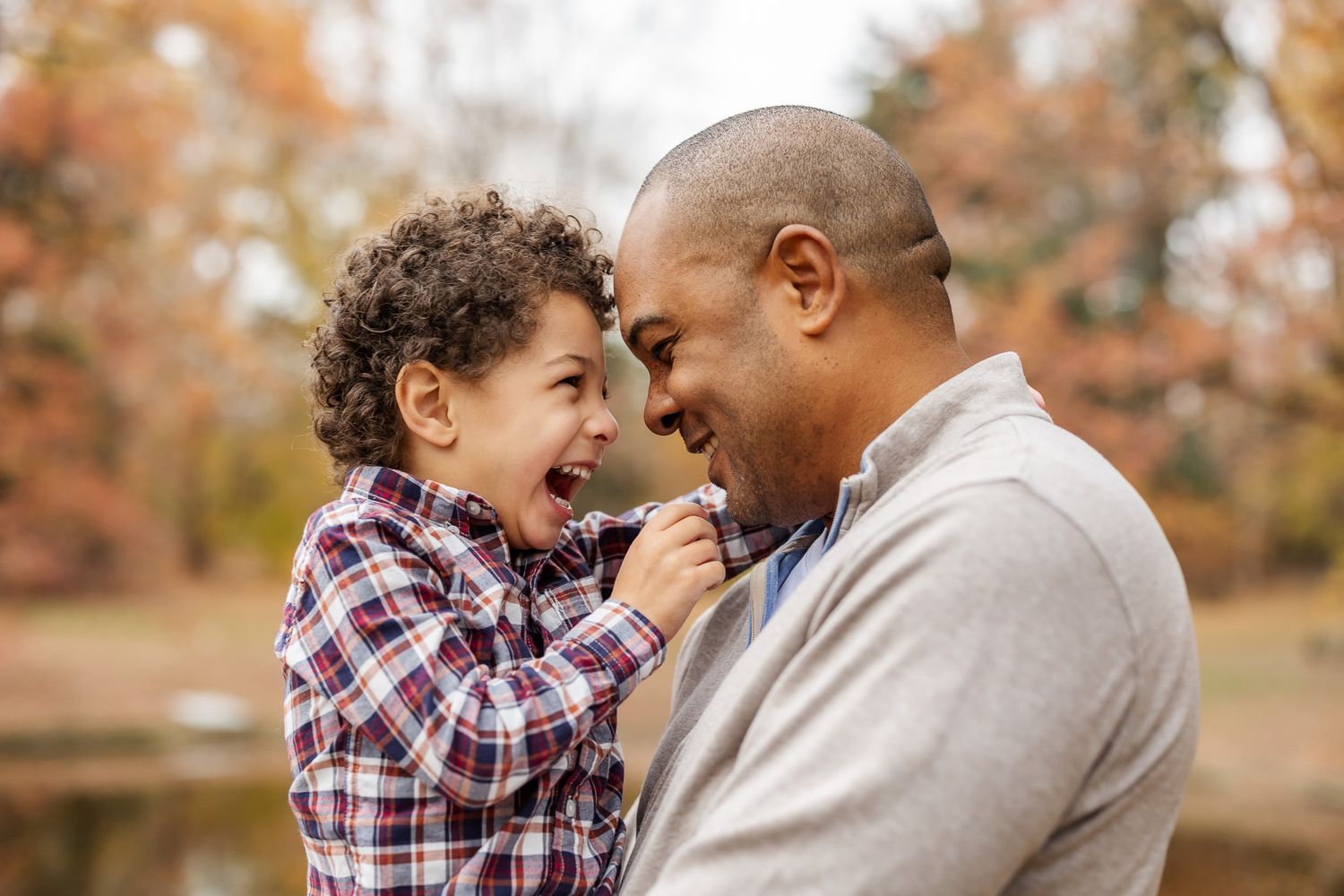 A joyful father and young son laugh together outdoors in an autumn park setting.
