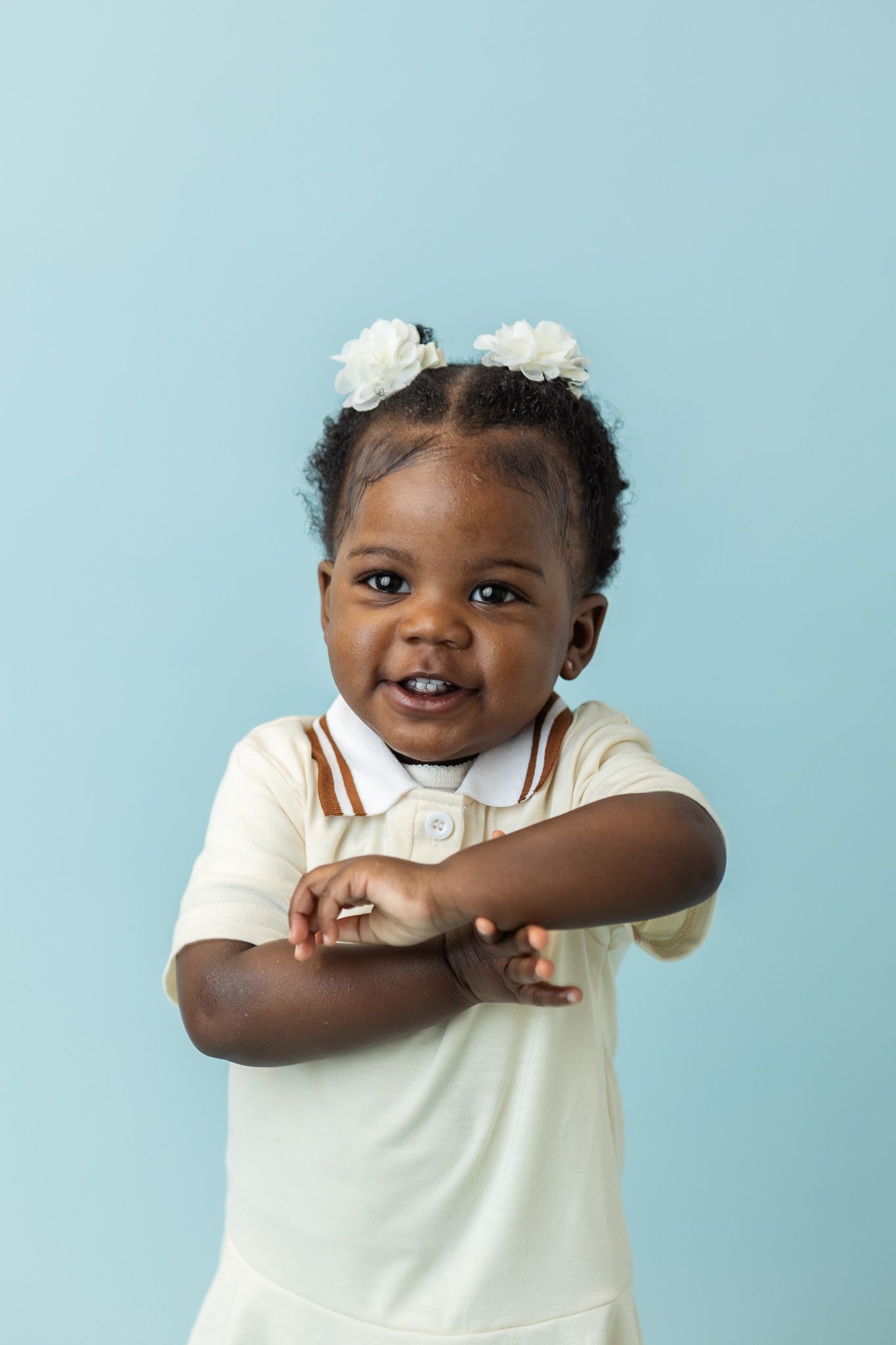 Adorable toddler girl with white flower hair accessories wearing cream polo dress, smiling against light blue background.