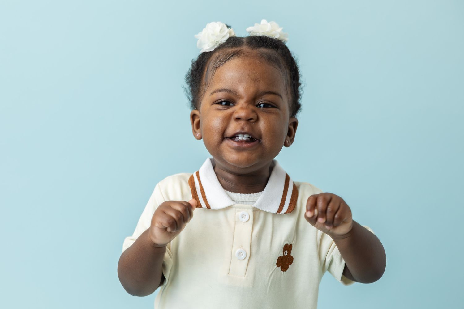 A happy toddler girl with white bows in her hair wearing a cream polo shirt, smiling against a light blue background.