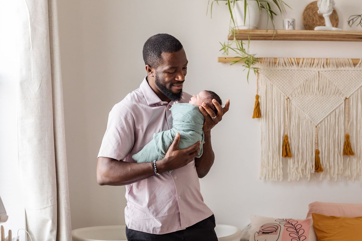 Parent holding and smiling at baby in light blue outfit against wall with macrame decor.