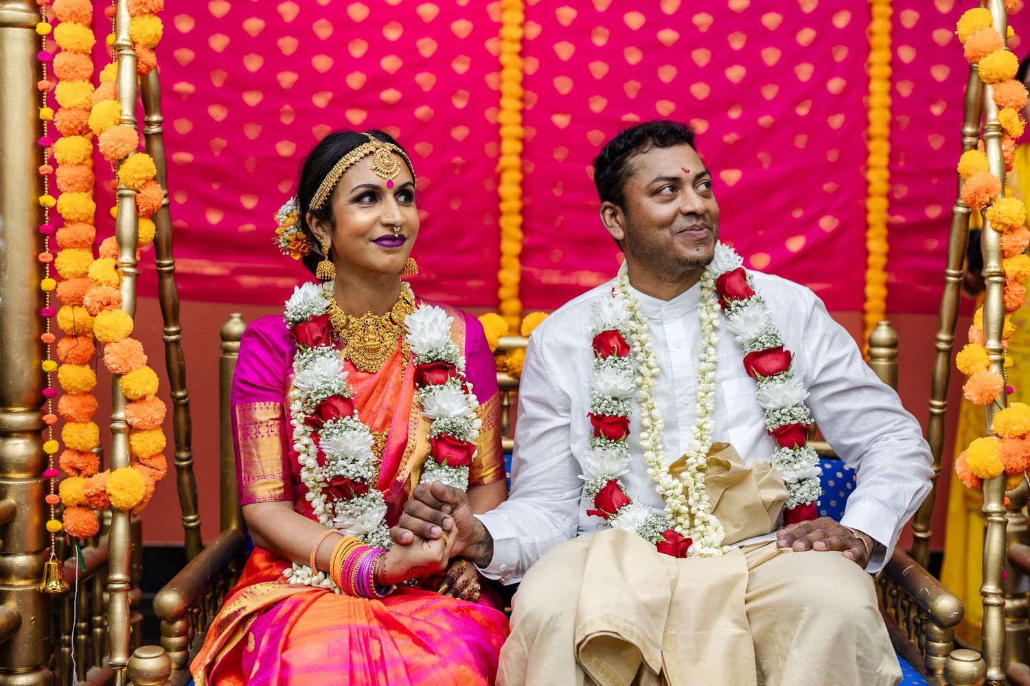 A traditional Indian wedding ceremony with people in white and gold attire seated under bright orange and pink decorations.