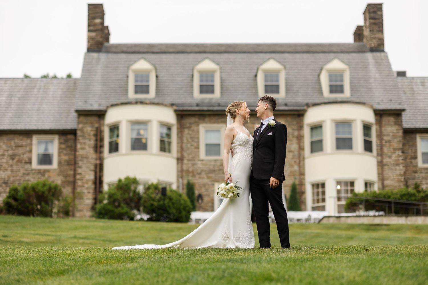 Bride and groom share a kiss on green lawn before elegant stone manor house on their wedding day.