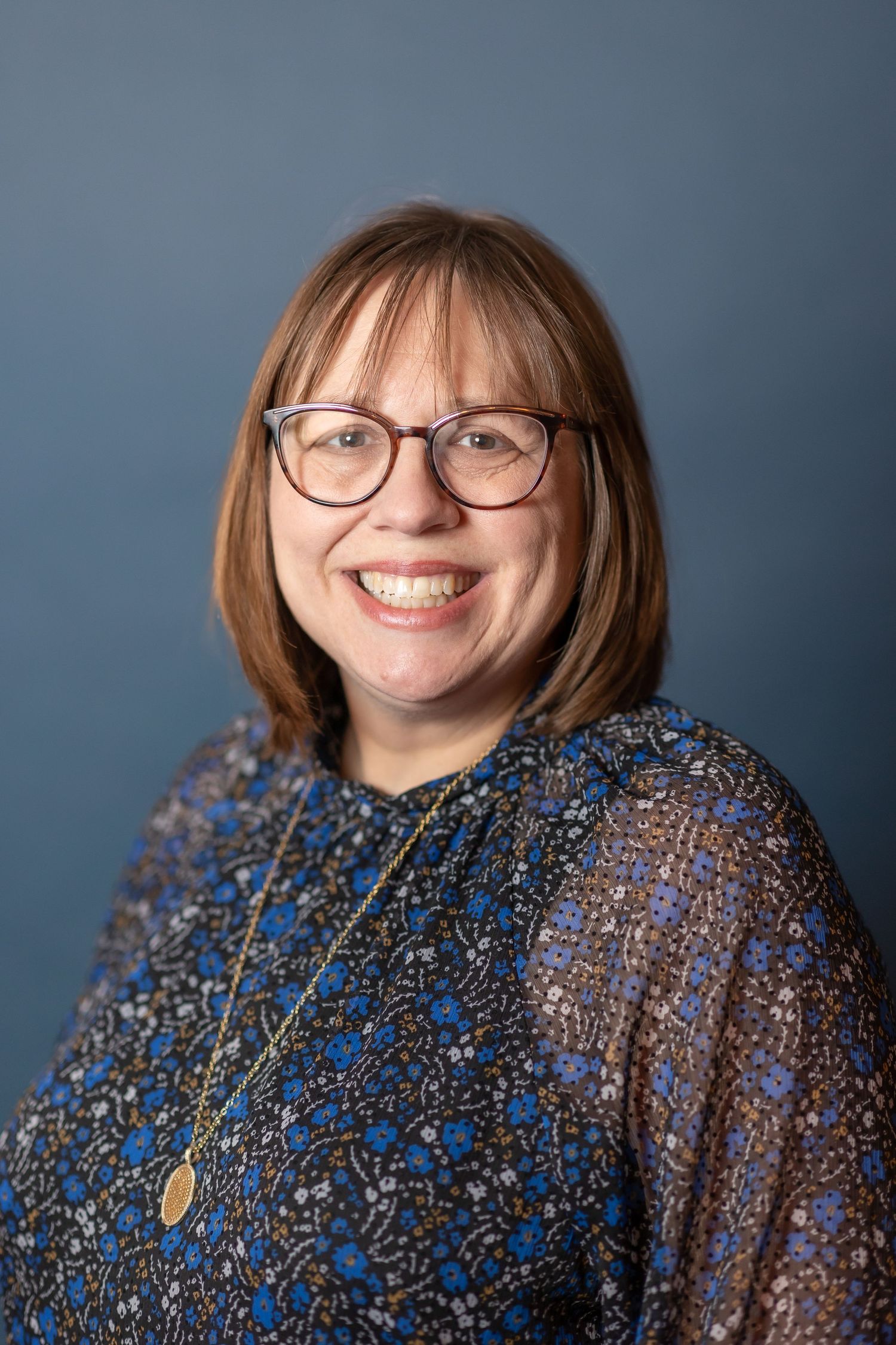 Welcoming professional portrait in patterned blue top against dark background.