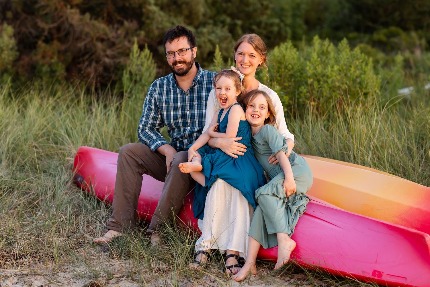 Family sits together on colorful surfboards at beach during sunset, surrounded by coastal grass.