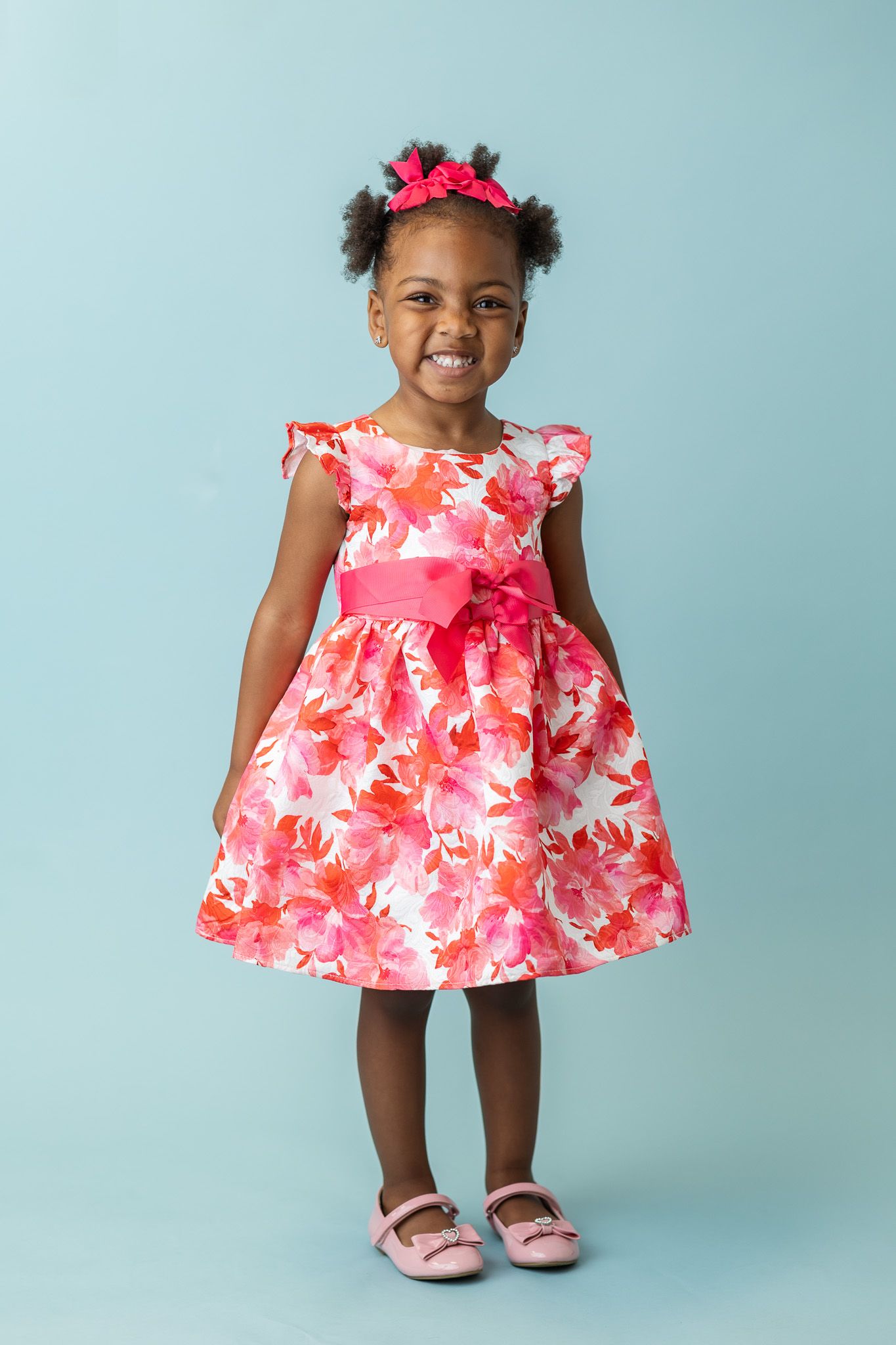Young girl in pink floral dress with bow, wearing pink hair accessories, smiling against blue background.