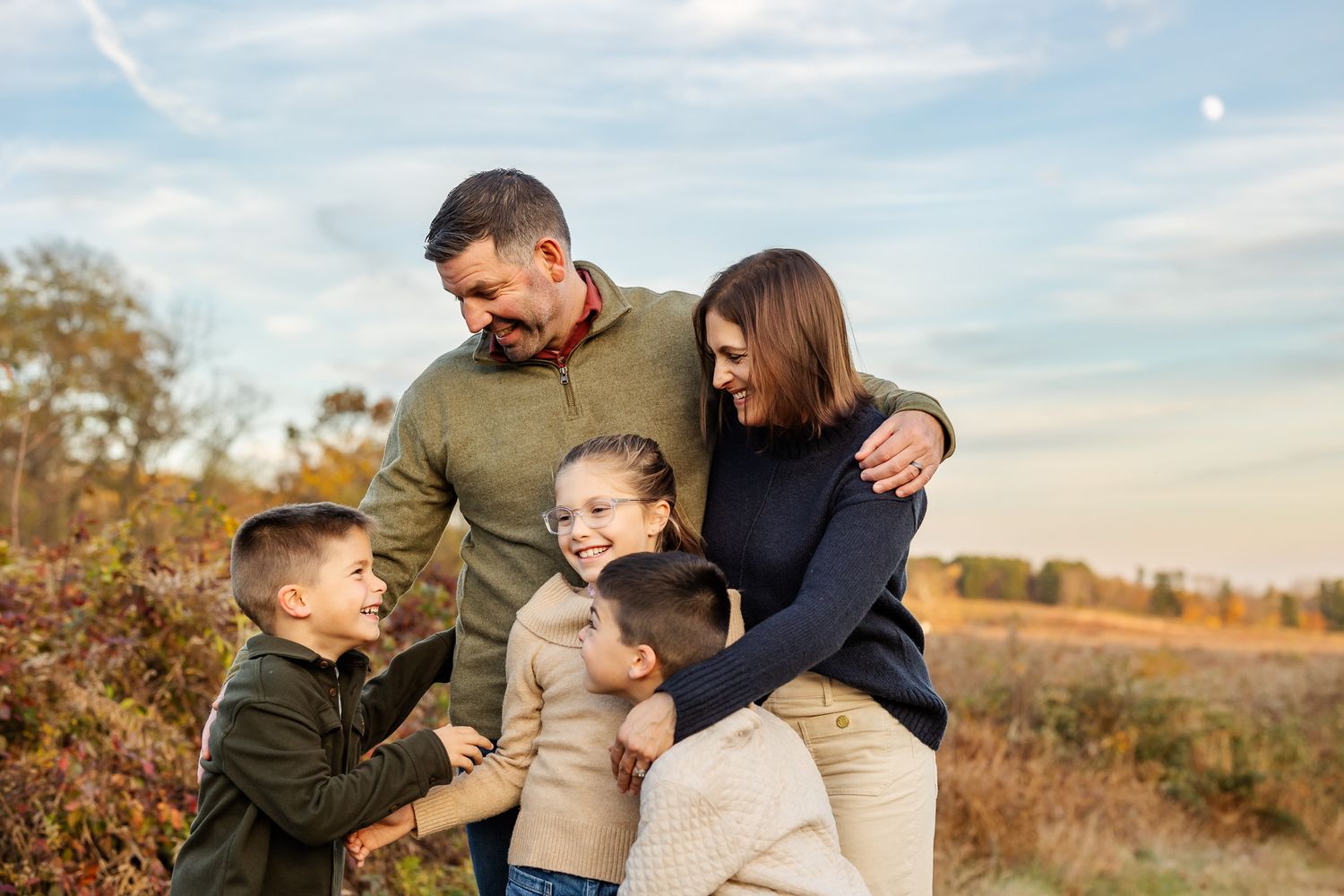 Happy family of five embracing outdoors in autumn field, parents hugging children in warm fall clothing under blue sky.