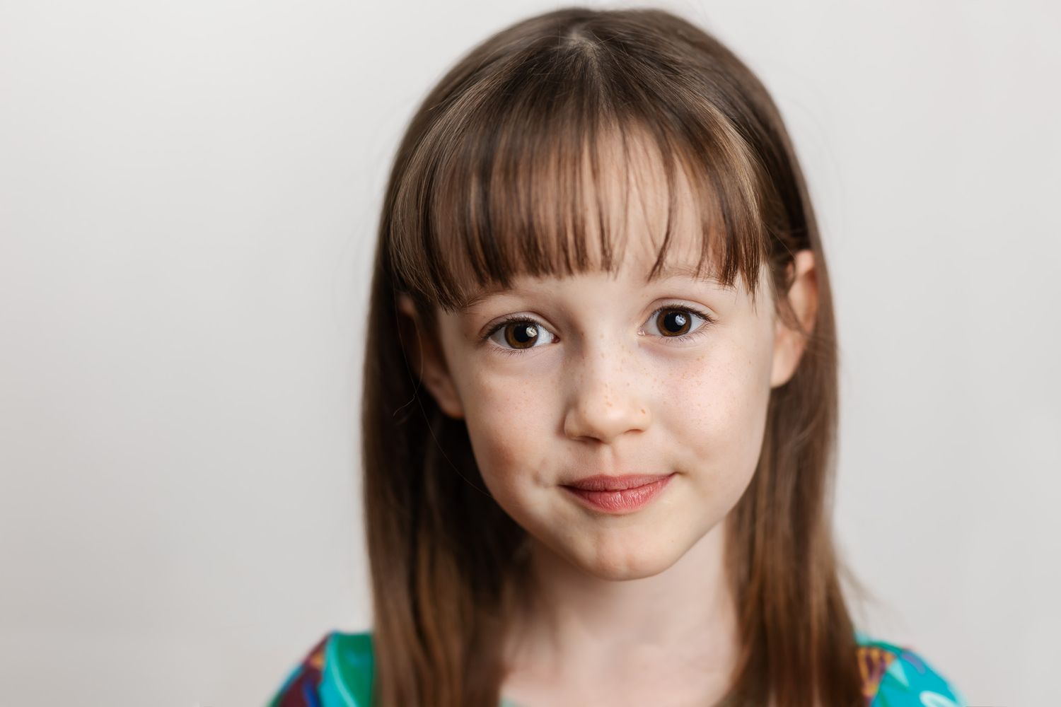 Portrait of a young girl with long brown hair and bangs, smiling softly against a white background.