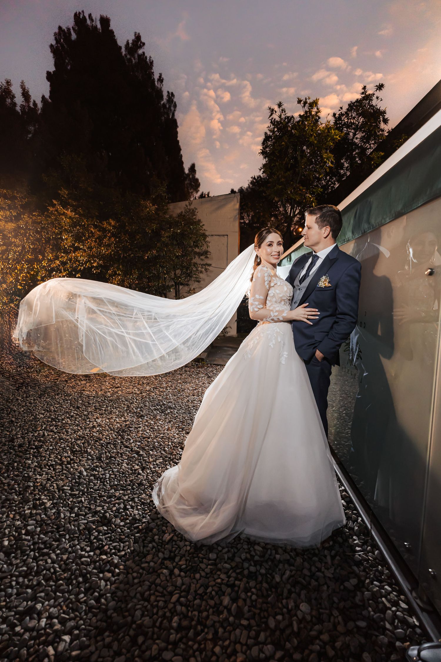 Novios se miran al aire libre de noche; velo largo de novia flota espectacularmente. Él traje azul. Fotografía de boda.