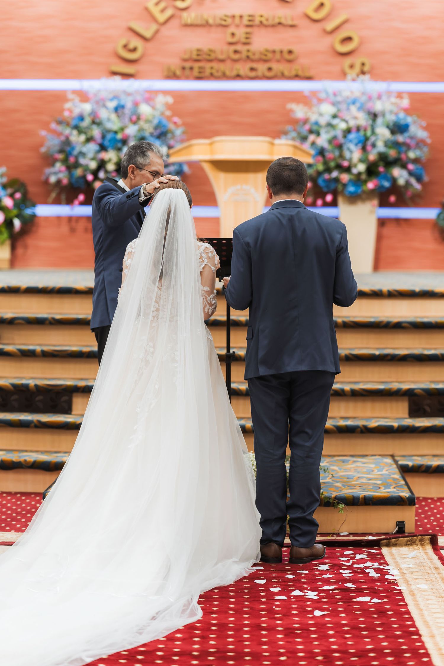 Vista trasera novios en altar durante ceremonia de boda IDMJI; oficiante junto a ellos. Novia velo largo. Iglesia .