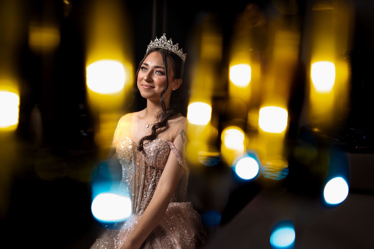 Retrato artístico: quinceañera sonriente con vestido rosa y tiara rodeada de luces bokeh amarillas y azules desenfocadas.