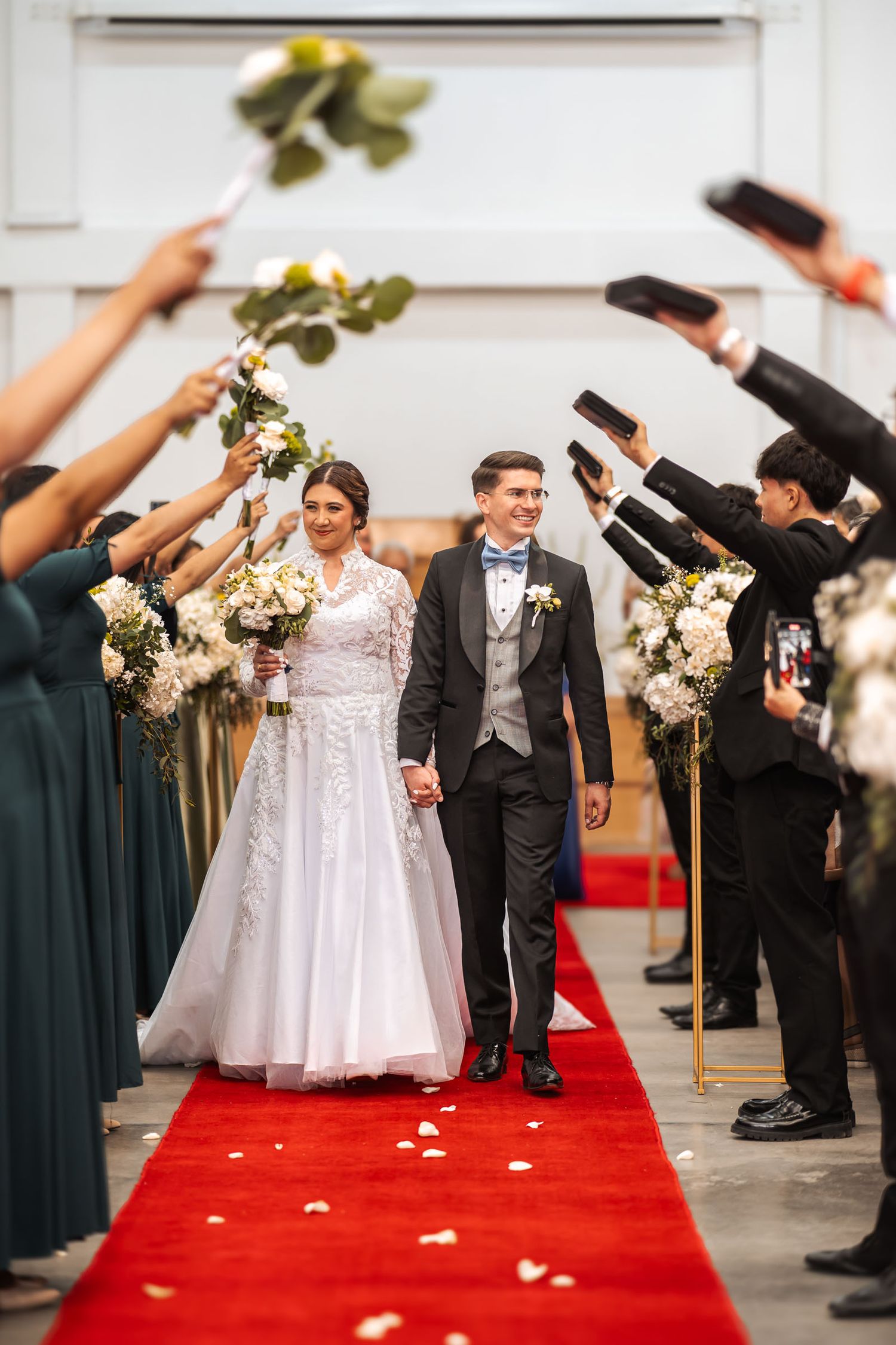 Pareja de novios desfila por pasillo de iglesia con alfombra roja, cortejo celebra levantando brazos y flores. Boda IDMJI