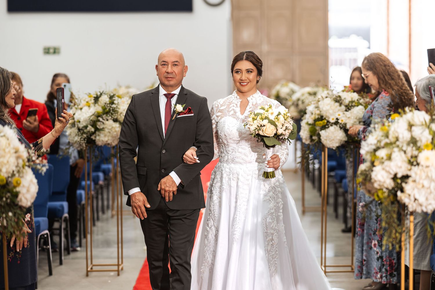 Novia con su padre por pasillo rojo de iglesia durante su entrada en ceremonia de boda IDMJI. Ramo