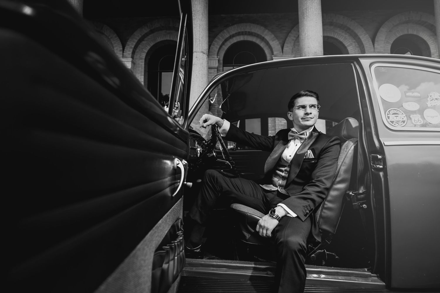 oto blanco y negro: Novio con esmoquin sentado elegante al volante de VW Escarabajo clásico con puerta abierta. Boda.