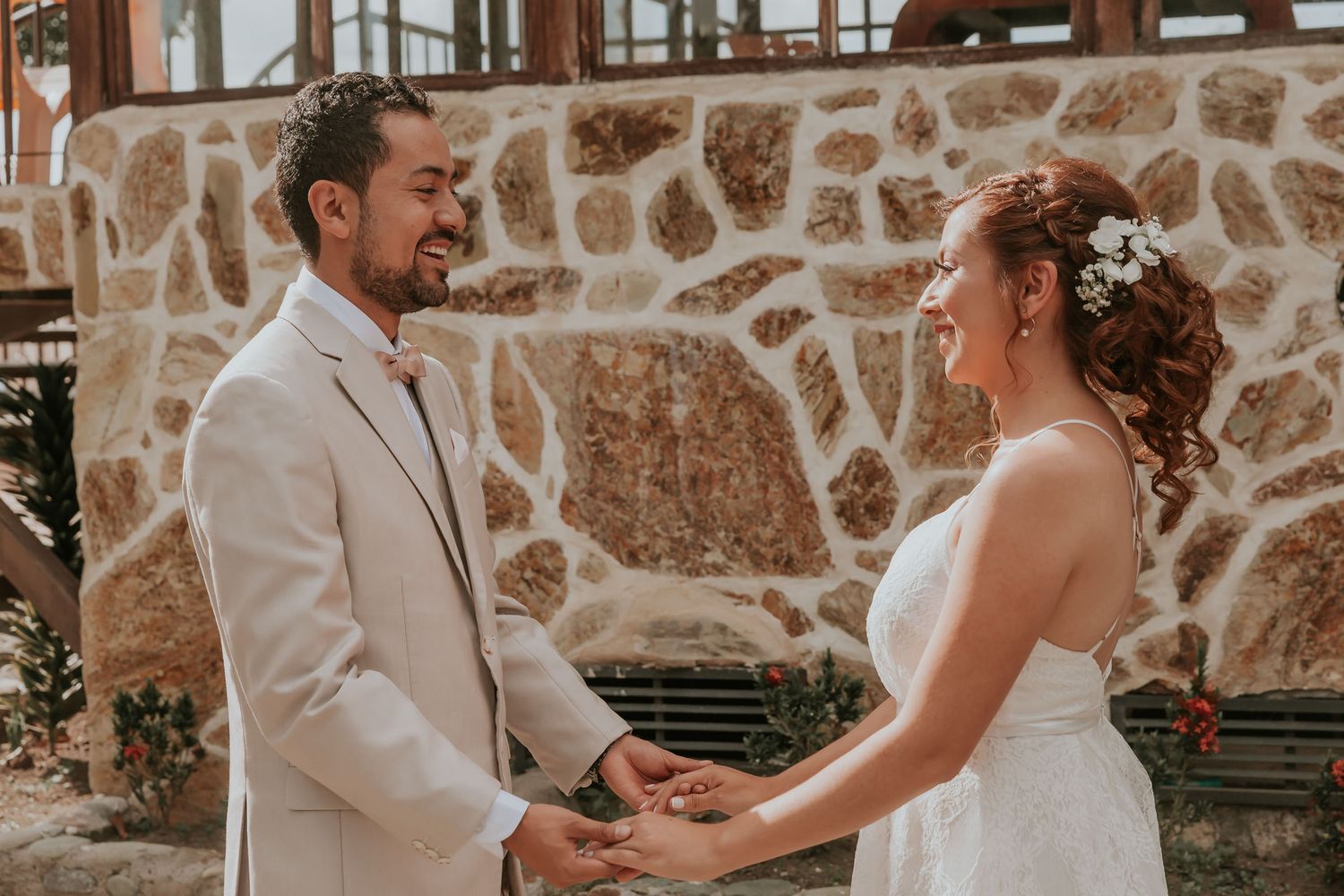 Novios sonrientes se toman de las manos mirándose; 'first look' boda frente a muro de piedra rústico.