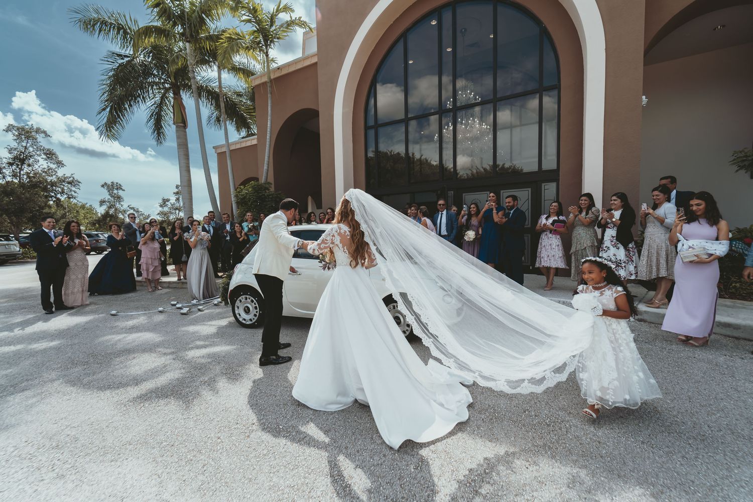 Novia con velo largo baja de coche blanco; novio ayuda, niña flores observa. Invitados, palmeras fondo.