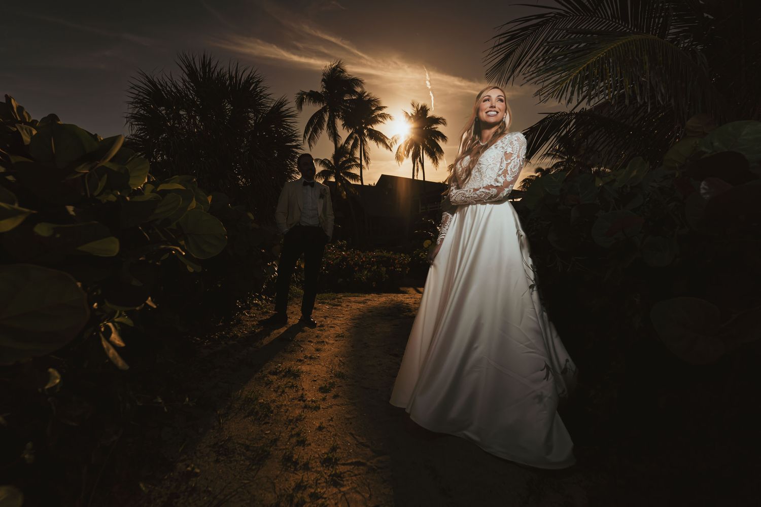 Novia ríe feliz de espaldas al atardecer dorado; novio silueta fondo. Palmeras, boda romántica.
