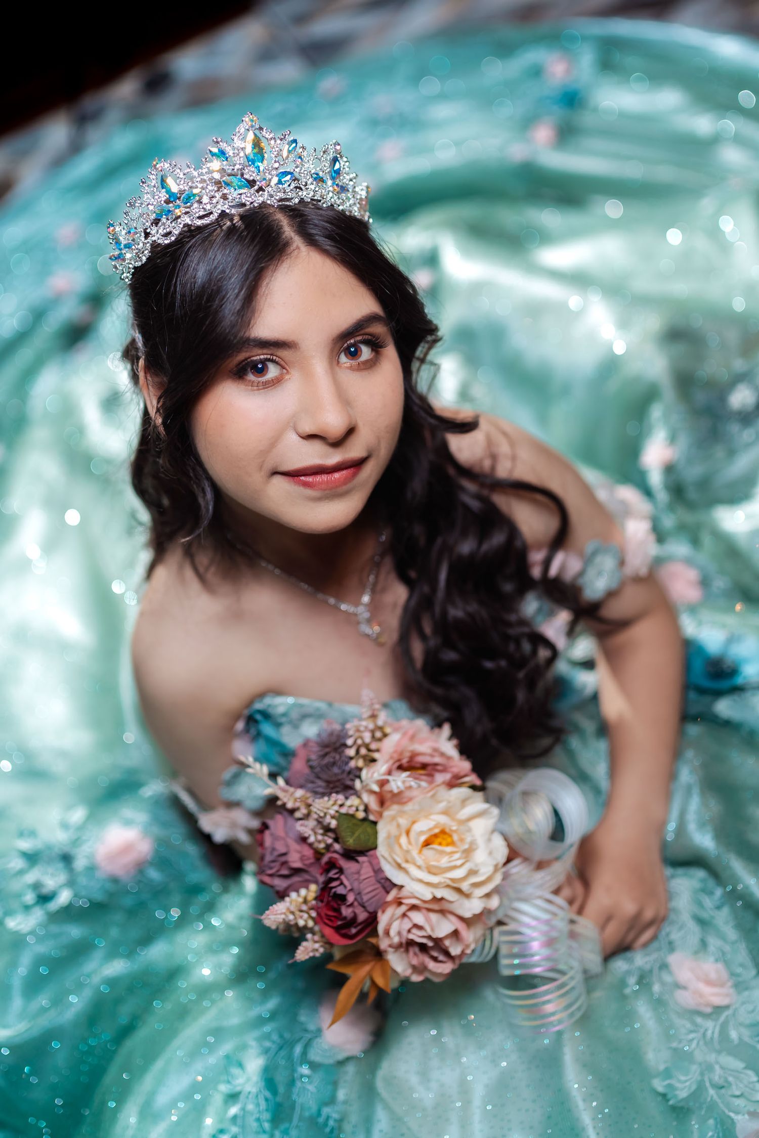 Primer plano cenital quinceañera con tiara y ramo, mira a cámara; vestido aguamarina, pétalos alrededor.