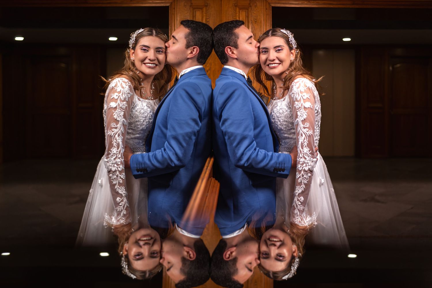 Foto boda artística: Novios sonrientes reflejados creando simetría; él besa su sien. Efecto espejo.