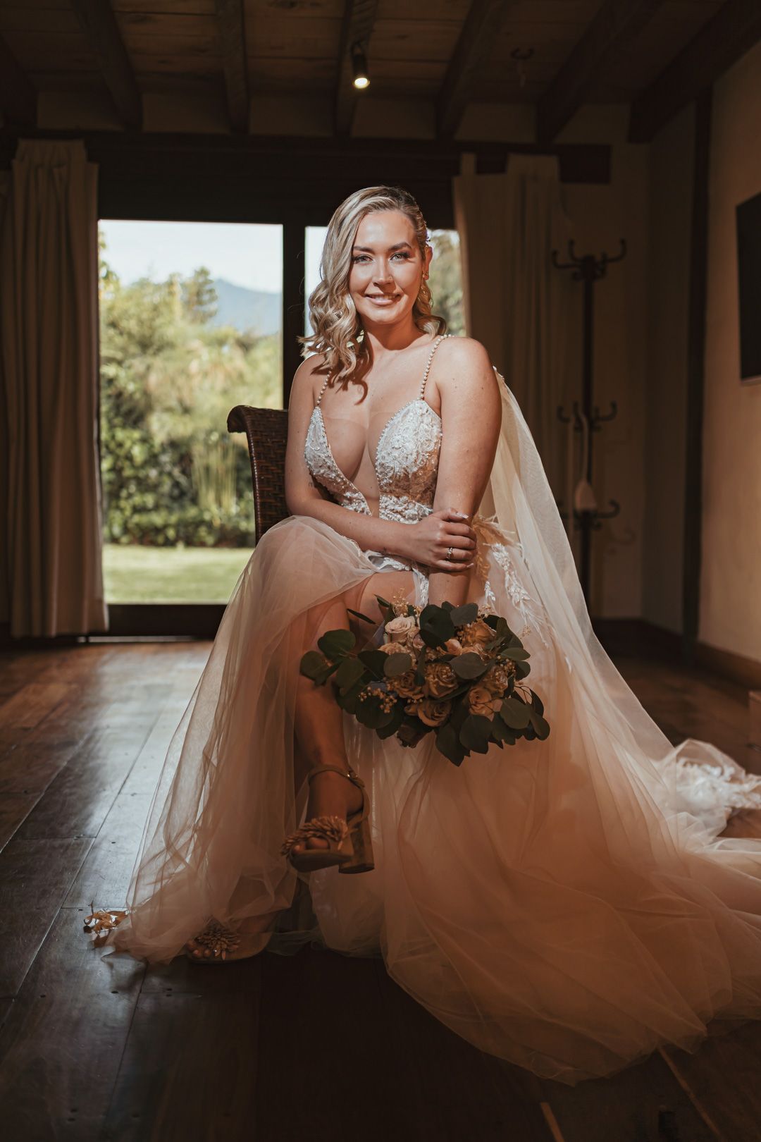 Sonriente novia rubia con ramo de flores y vestido blanco de encaje sentada en silla. Luz natural entrando por ventana con vista a paisaje