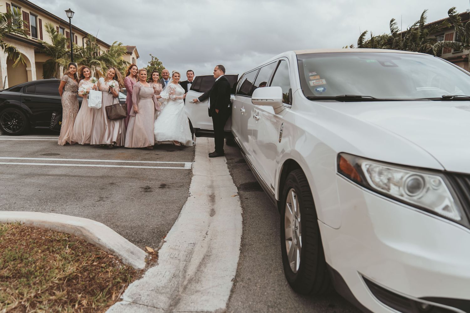 Grupo nupcial (novia, damas de honor) posa junto a limusina SUV blanca en exterior; chófer presente. Día boda Miami.