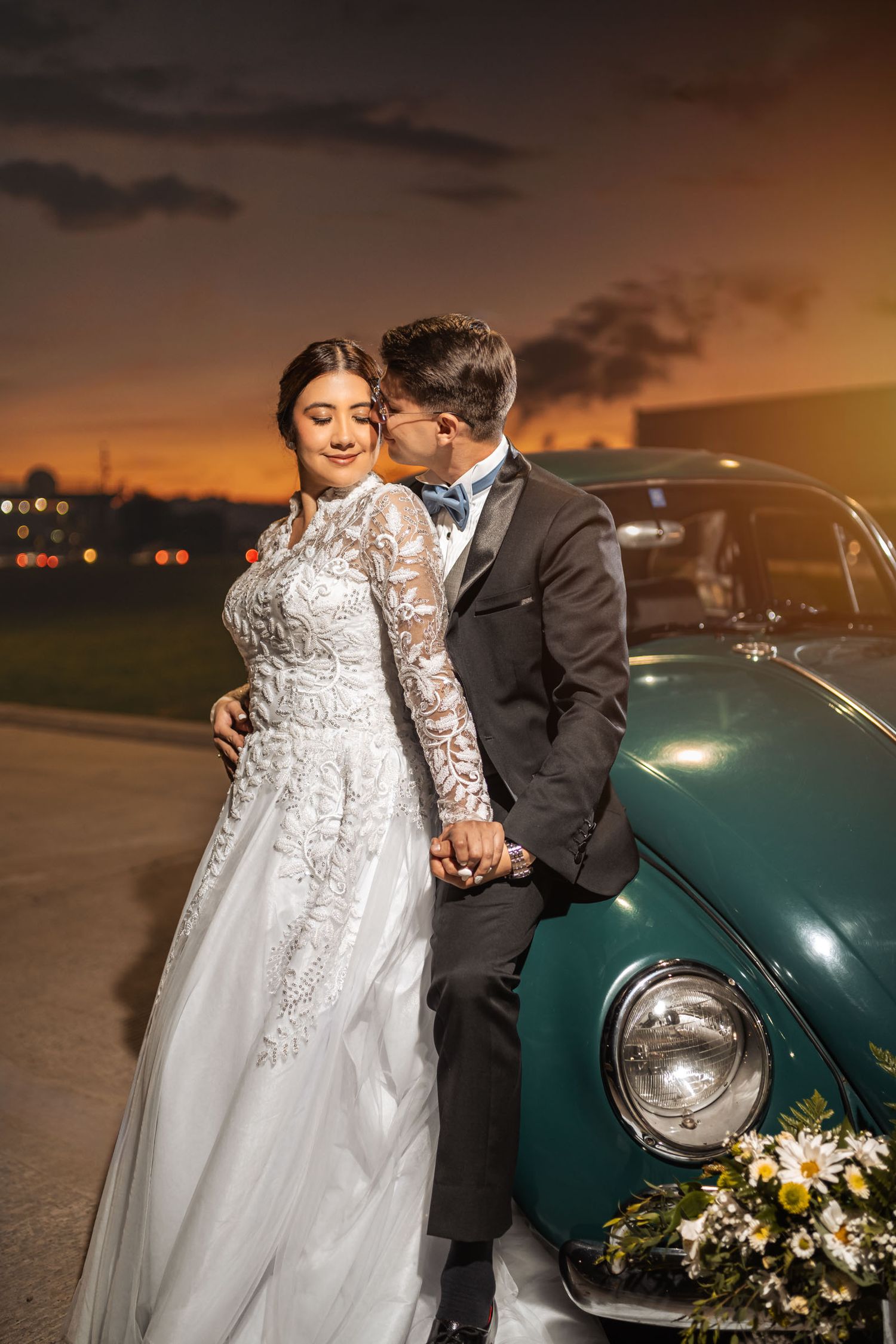 Novios tomados de la mano miran paisaje al atardecer junto a coche verde clásico. Abrazo tierno en boda.