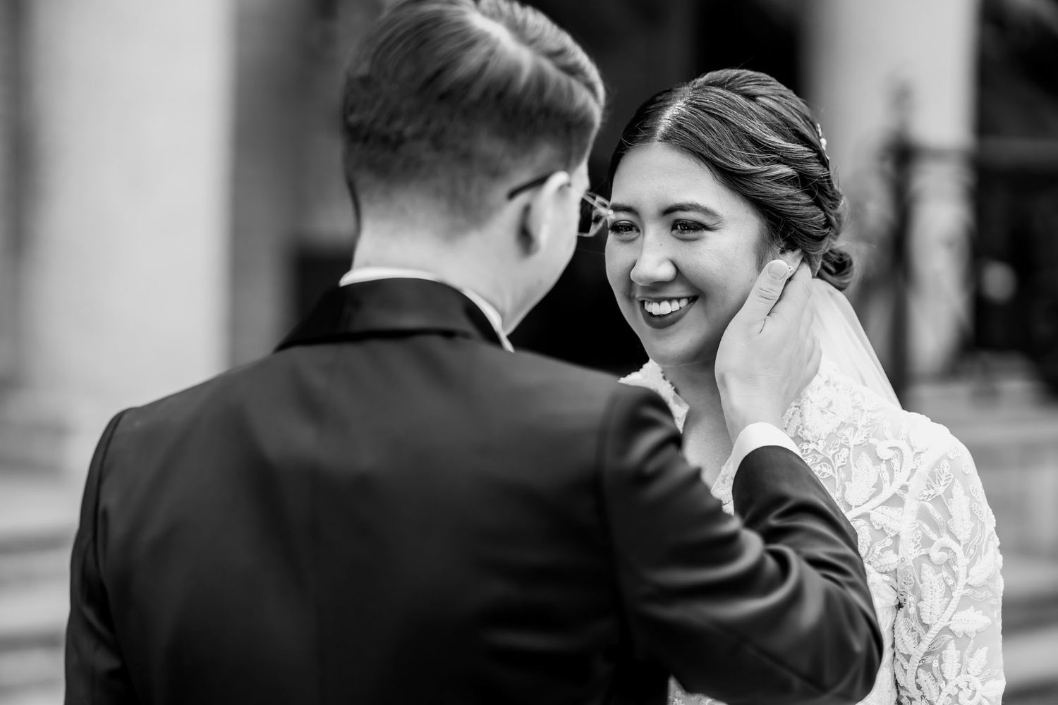 Foto blanco y negro: Novio acaricia mejilla de novia sonriente; momento tierno de boda visto sobre el hombro de él.