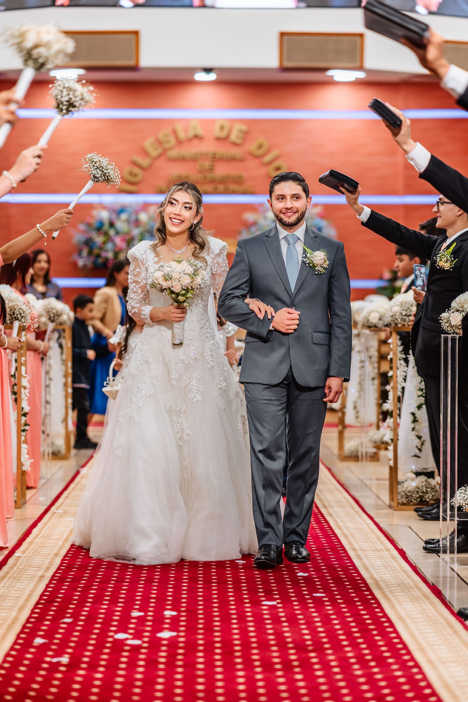 Novios caminan sonrientes por pasillo de alfombra roja, invitados celebran su paso levantando ramos y biblias. Boda idmji