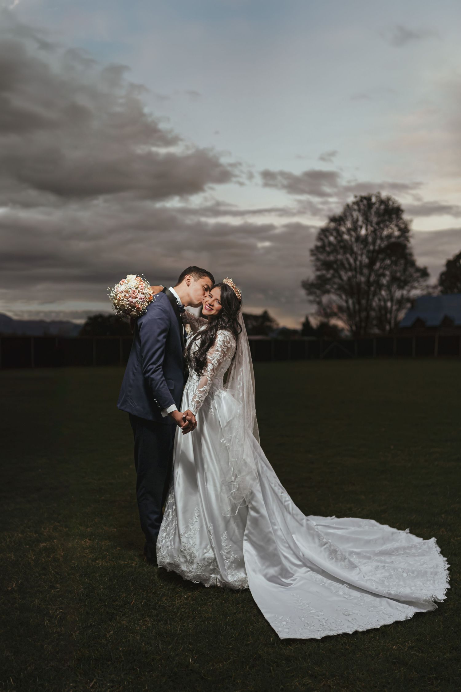 Novios se besan en campo al atardecer; novia con velo largo, cielo nublado. Foto boda romántica.