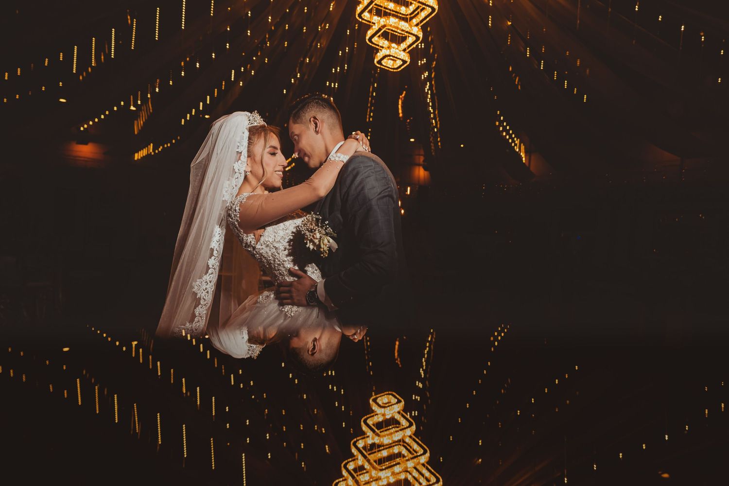 Novios se abrazan reflejados en piso brillante bajo líneas de luces cálidas; perspectiva. Boda romántica.