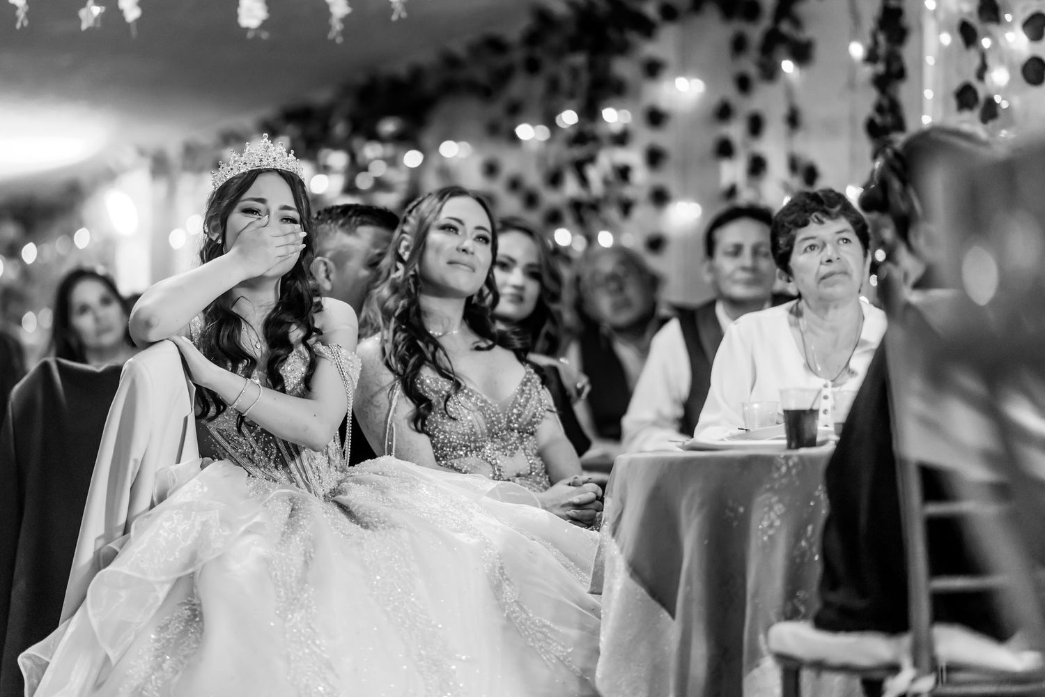 Foto B&W: Joven quinceañera reacciona emocionada cubriendo su boca en fiesta; sentada junto a otra joven.