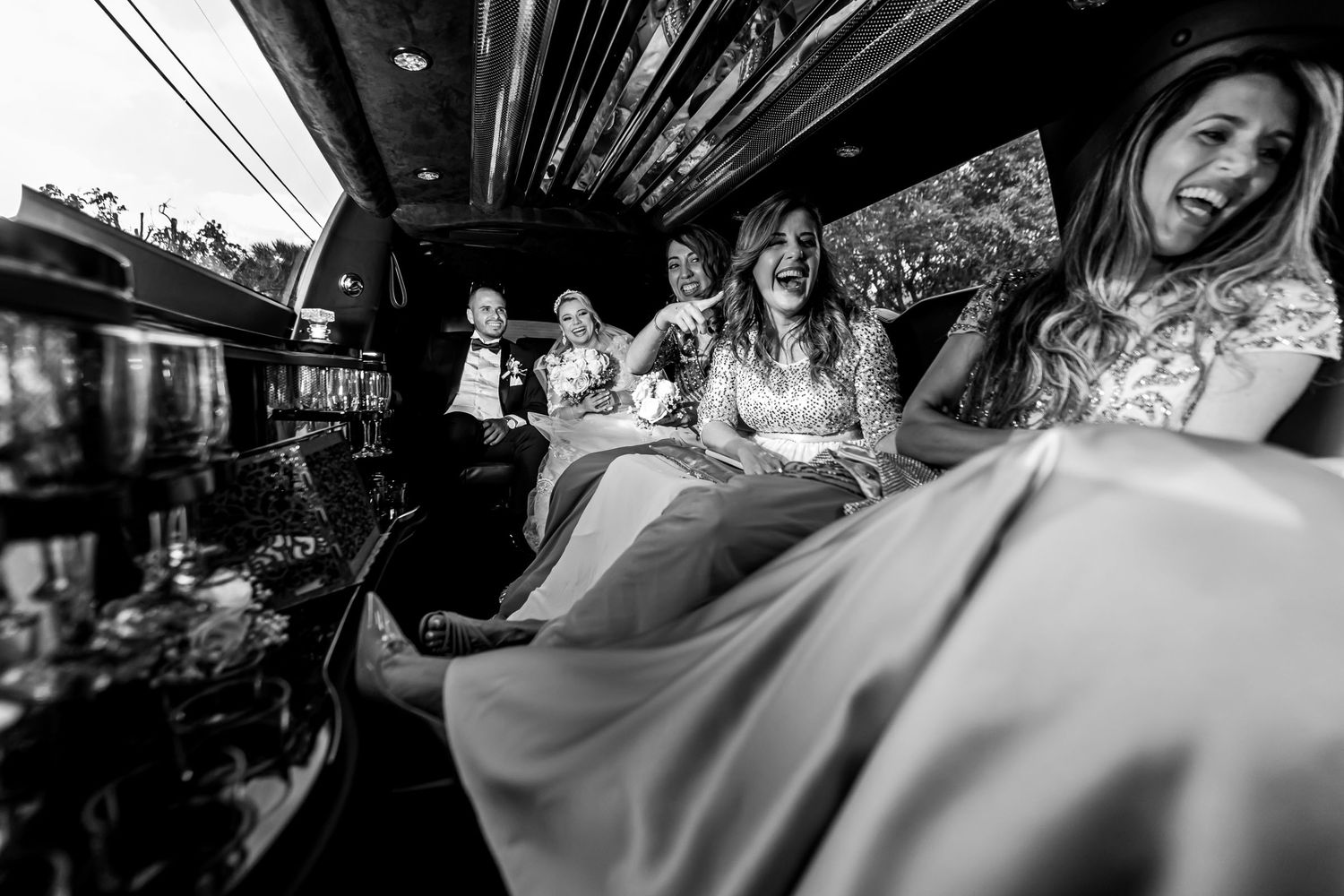 Foto B&W: Novia y damas de honor ríen a carcajadas dentro de limusina; momento divertido boda.