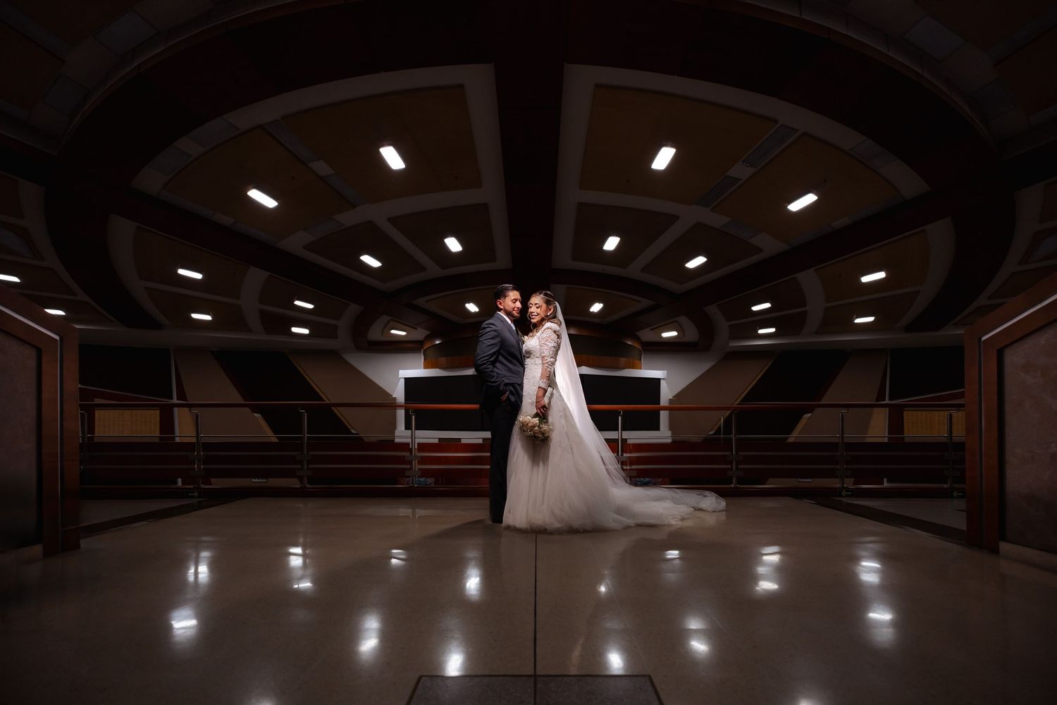 Novios se abrazan en un amplio salón interior con techo curvo e iluminación geométrica. Foto romántic de boda IDMJI en Colina Bogotá