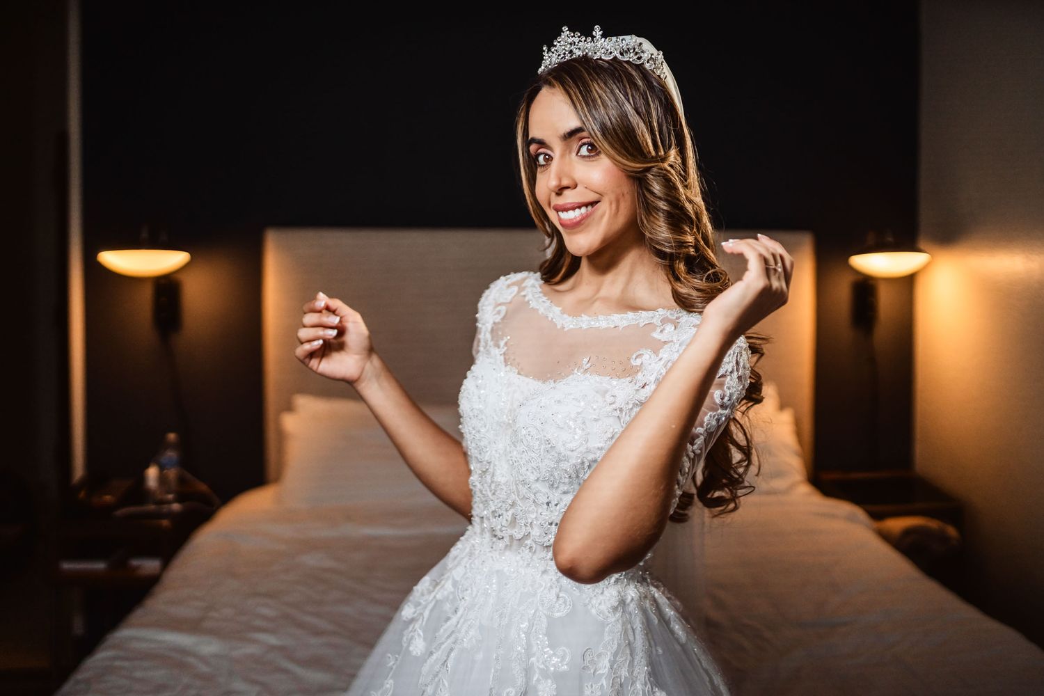 Novia sonriente con vestido blanco (mangas ilusión) y tiara posa sentada en cama; habitación hotel boda.