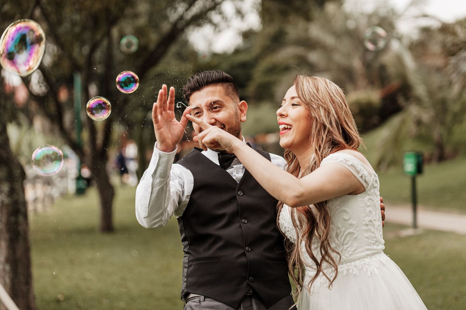 Novios ríen y juegan con burbujas de jabón en parque; momento divertido sesión fotos boda exterior.