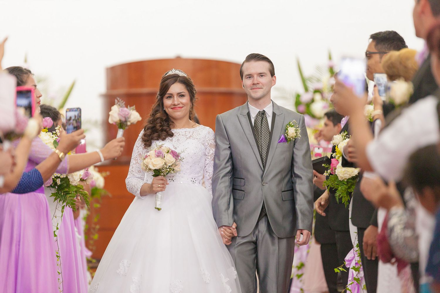 Pareja de novios caminando frente al altar, tomados de la mano y sonriendo, mientras todos los invitados los miran caminar la marcha nupcial
