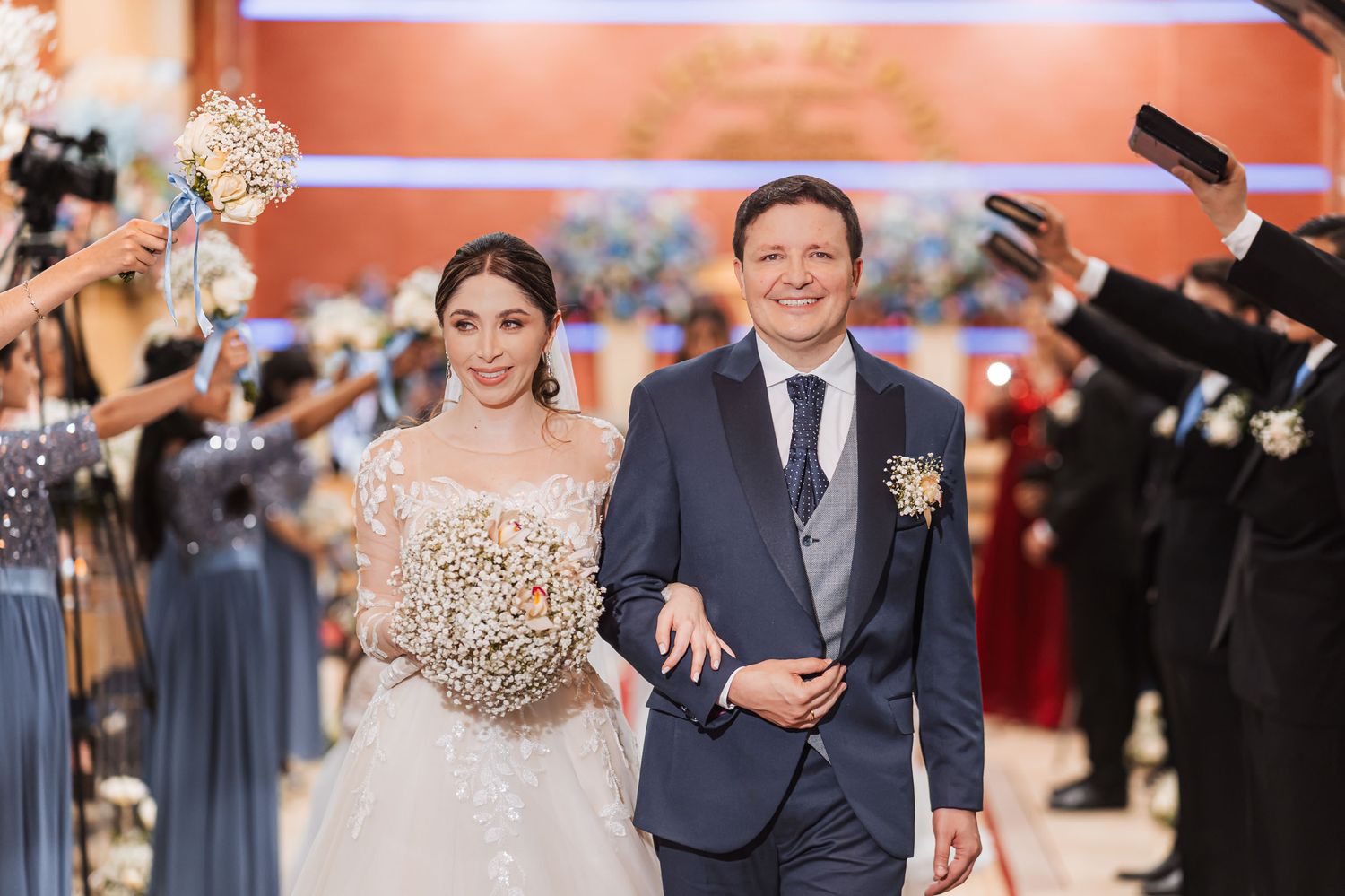 Novios sonrientes en salida nupcial por iglesia; cortejo con vestidos azules forma arco. Novia ramo gypsophila. Boda IDMJI