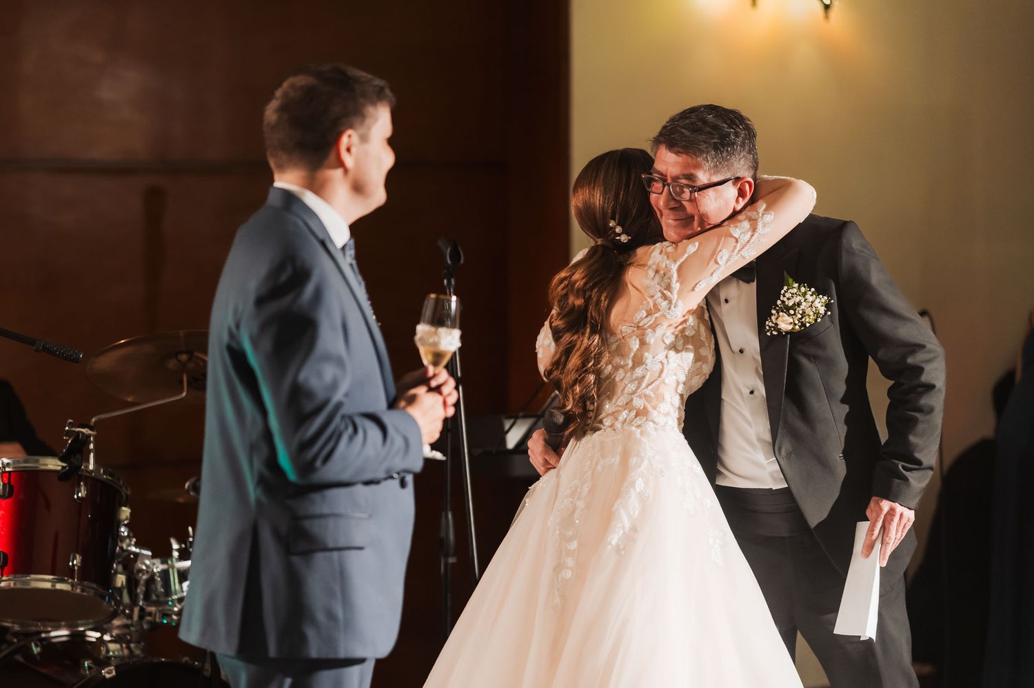Recién casados se abrazan emotivamente durante un brindis o discurso en su recepción de boda, invitados observan