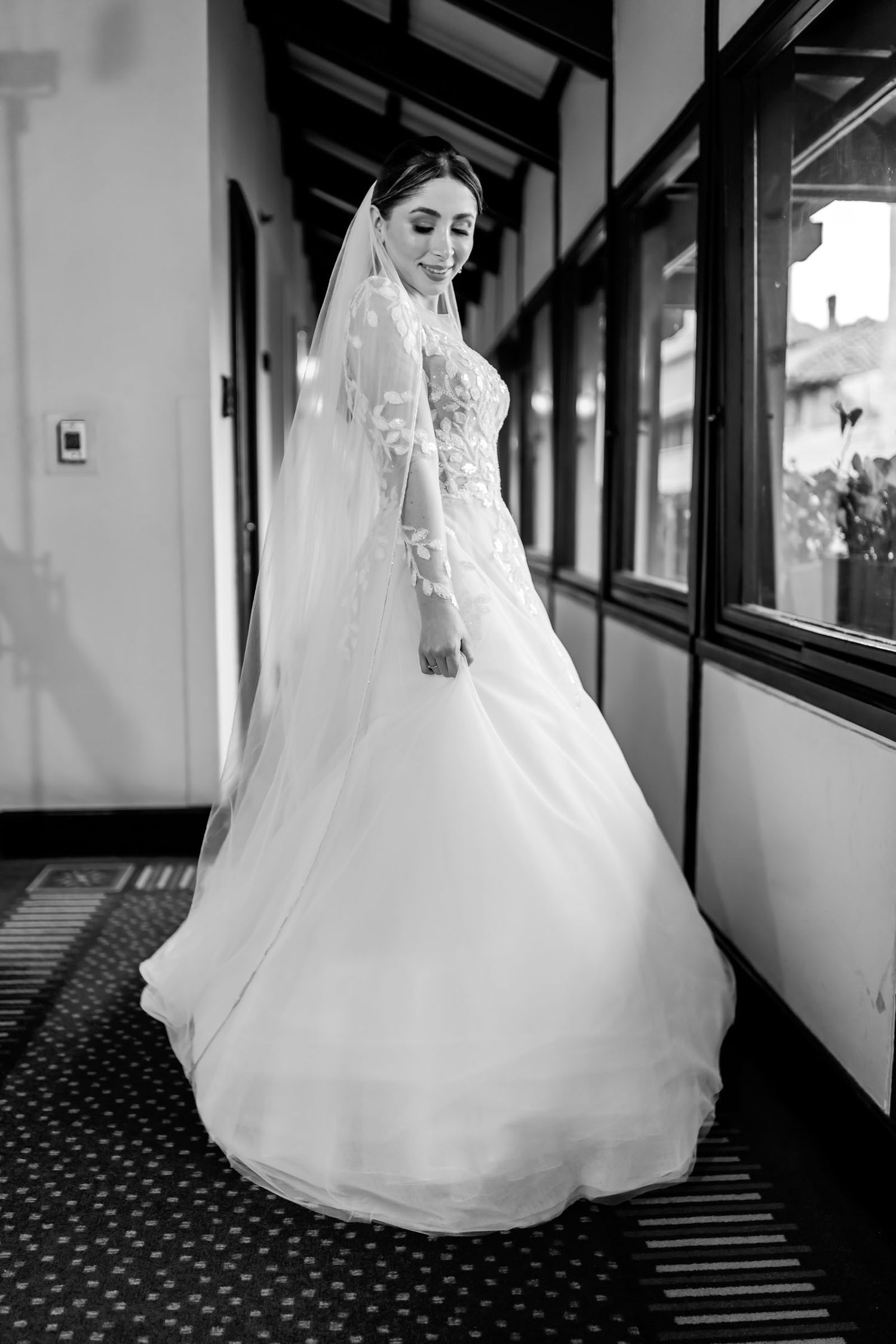 Foto B&W: Novia con vestido bordado y velo mira sobre el hombro en pasillo interior. Fotografía boda.
