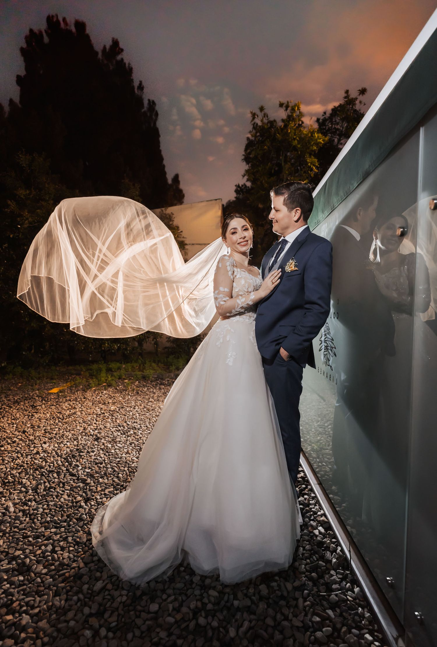 Novios posan de noche al aire libre; velo largo iluminado flota tras novia con vestido manga larga. Él traje azul. Boda.