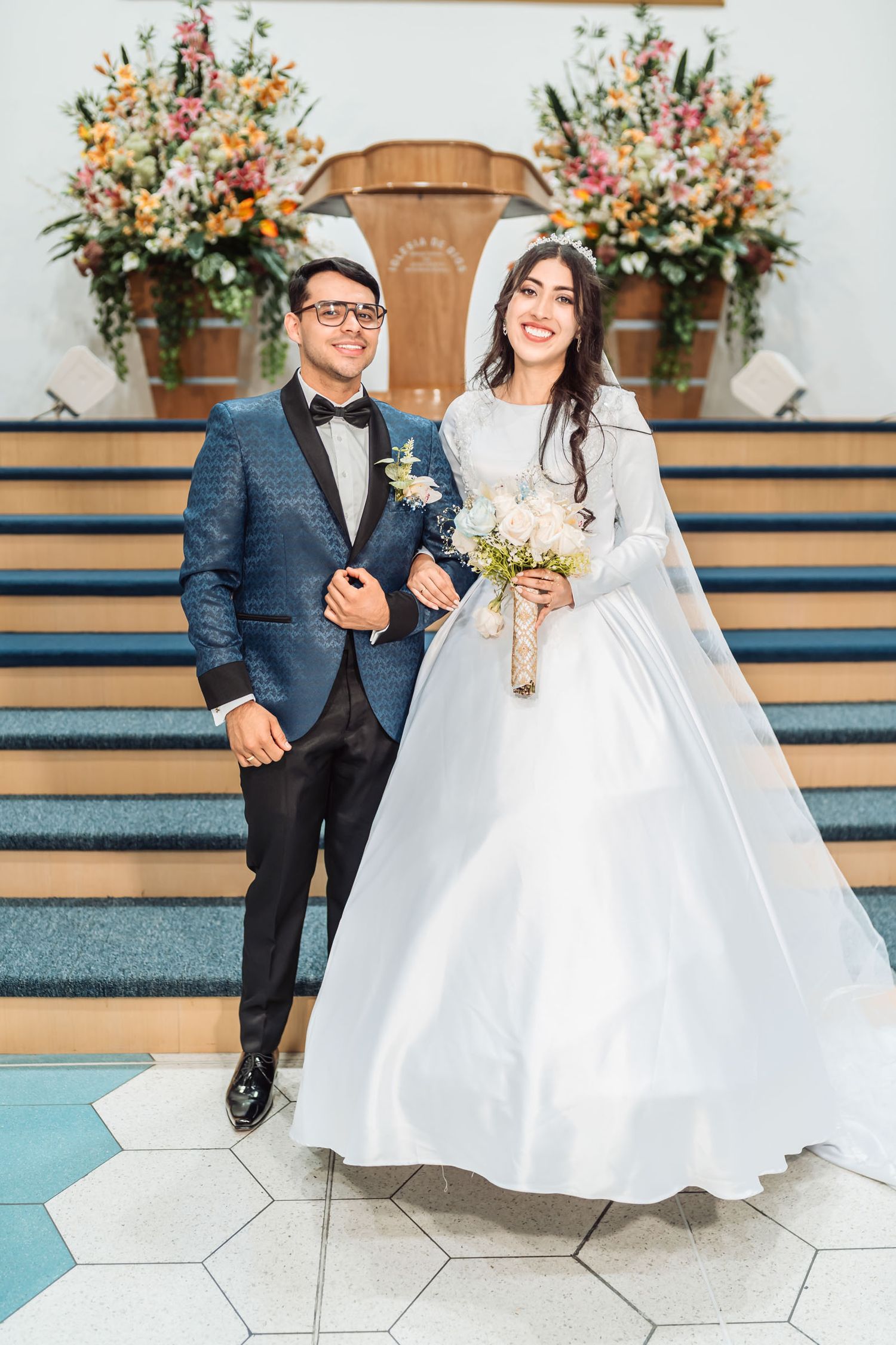 Novios sonrientes en altar iglesia; él traje azul brillante, ella vestido blanco, velo y ramo. Boda IDMJI.